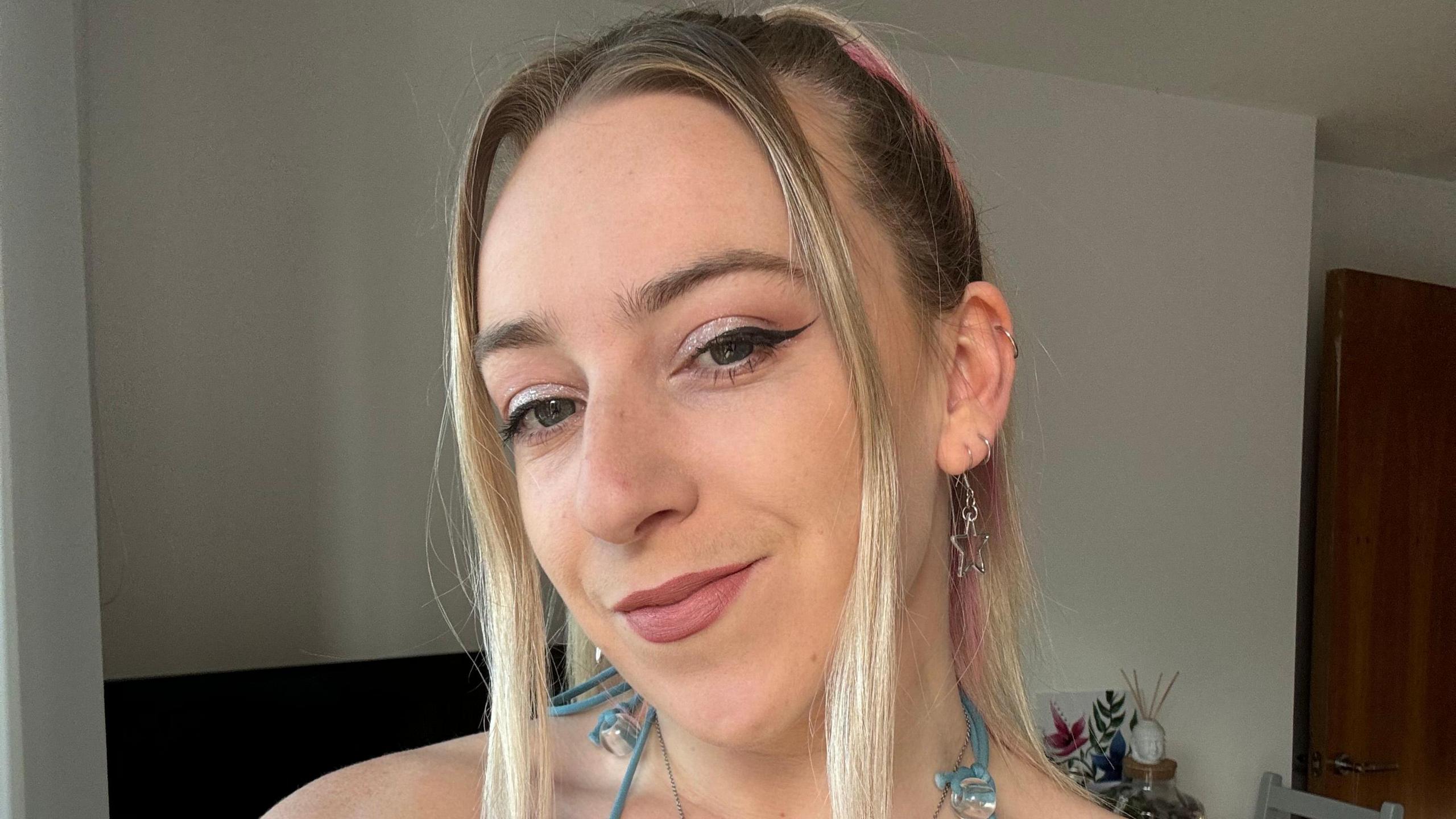 Abi smiles at the camera, her long blonde hair is pulled back from her face with strands at the front. She wears silver earrings and sparkly eyeshadow on her eyes.