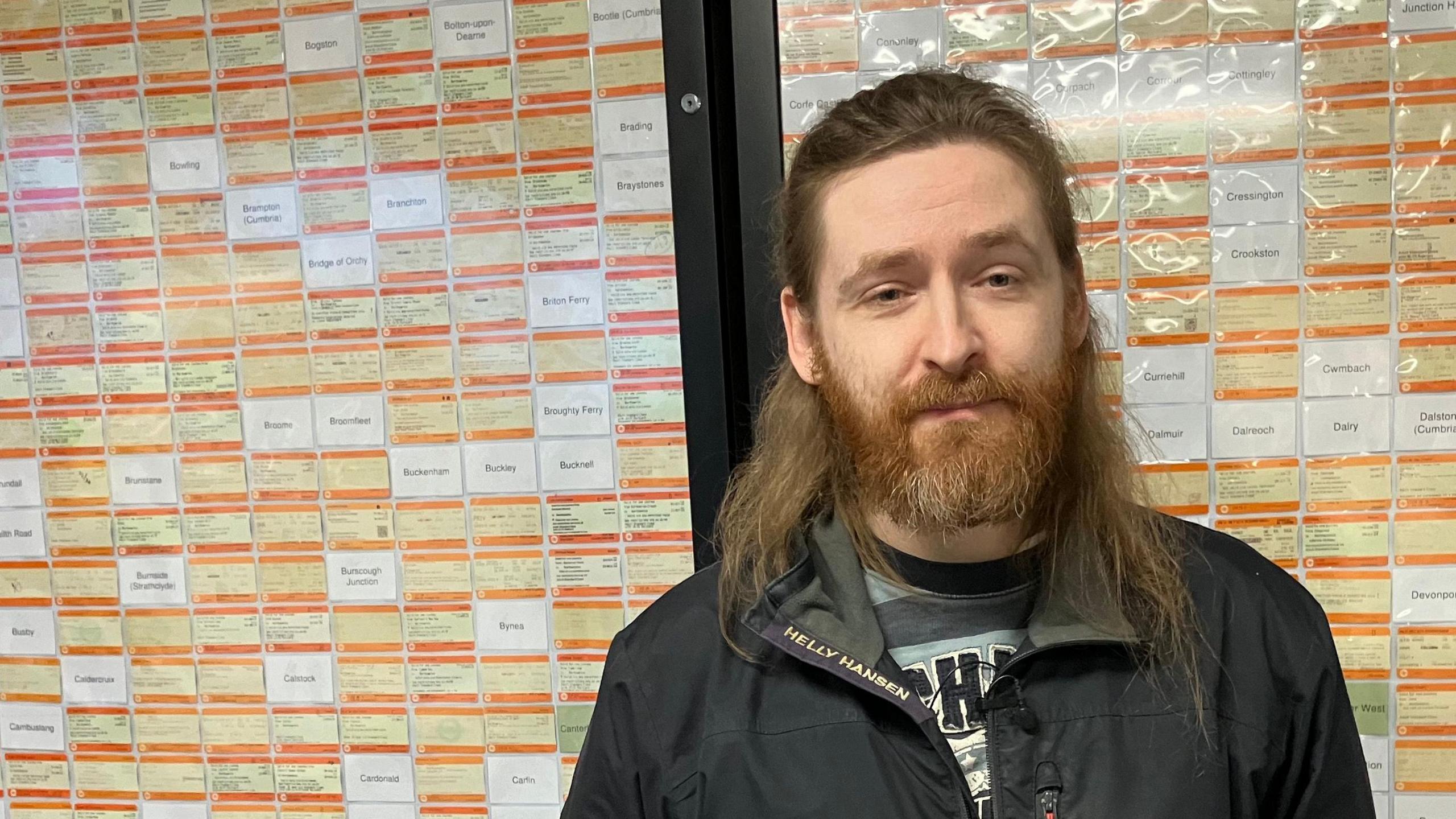 Elliott Badger who has should-length brown hair pulled back from his forehead and a beard. He is wearing a black jacket and standing in front of a notice board which is packed with railway tickets. There are also white cards with the type-written names of missing stations