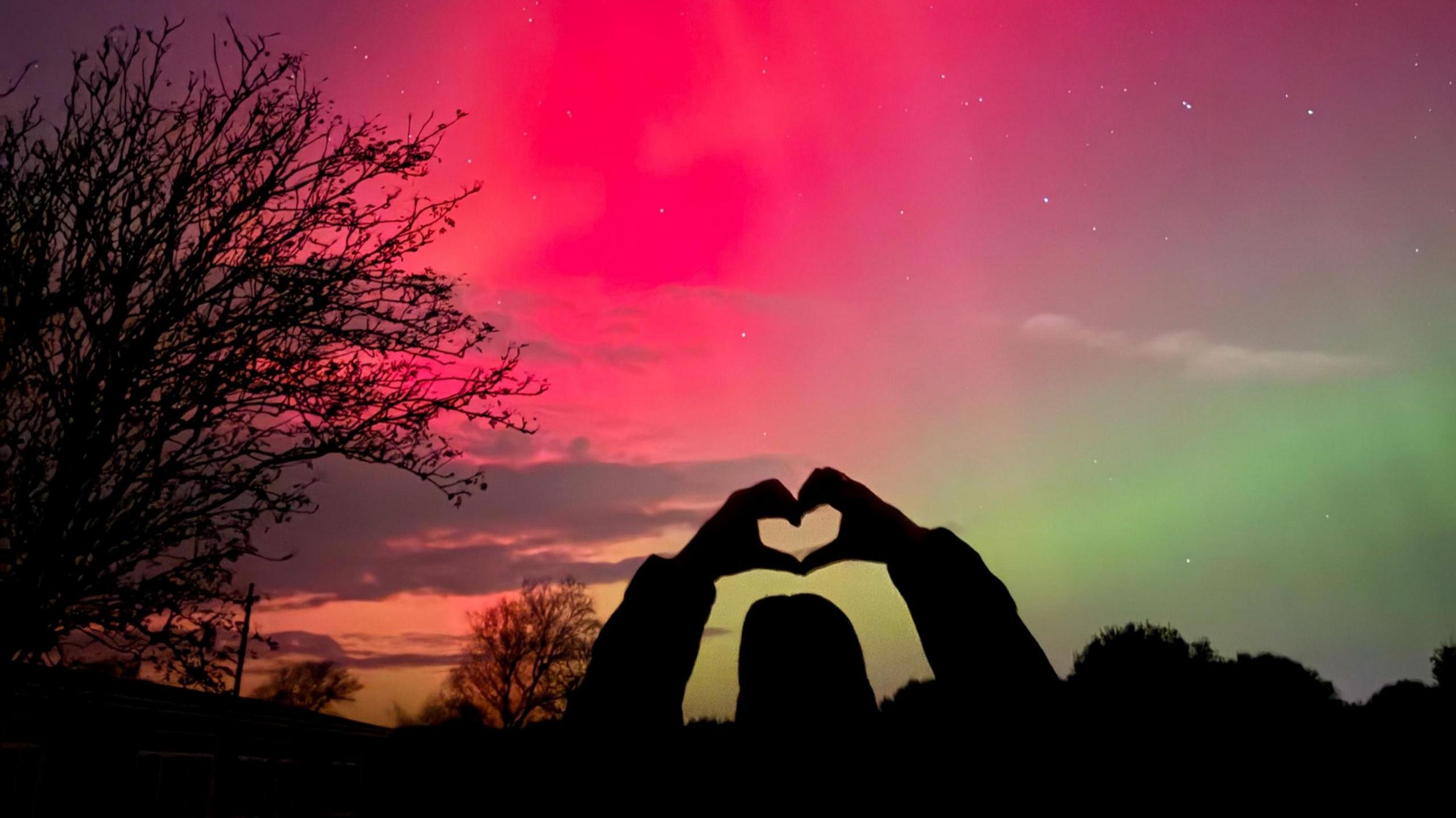 A silhouette of a person holding up a love heart directly towards a backdrop of the Northern Lights in bright red, bright green and darker orange and purple tones