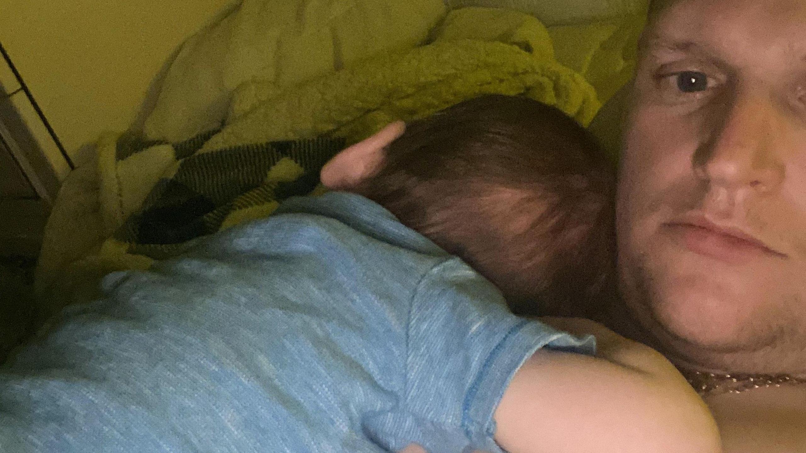 A young boy in a blue shirt lies asleep on the chest of man taking a selfie. He has a gold chain around his neck.