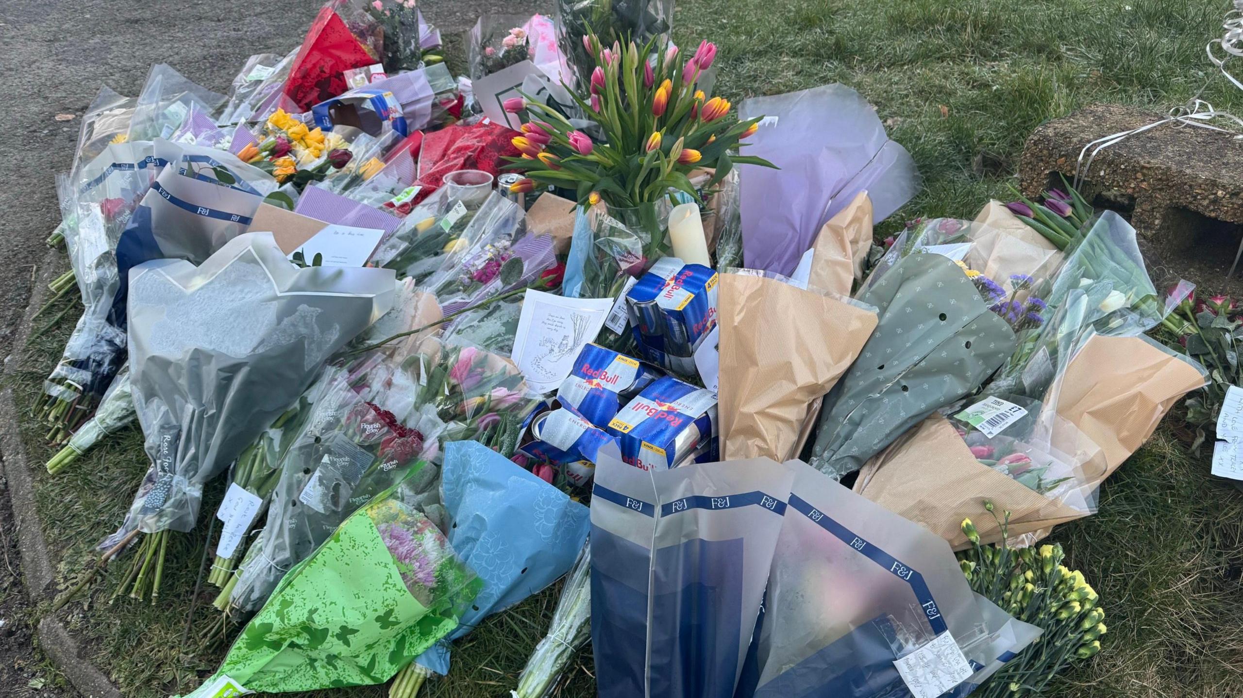 A collection of flowers laid on the ground around some boxes of Red Bull energy drinks.