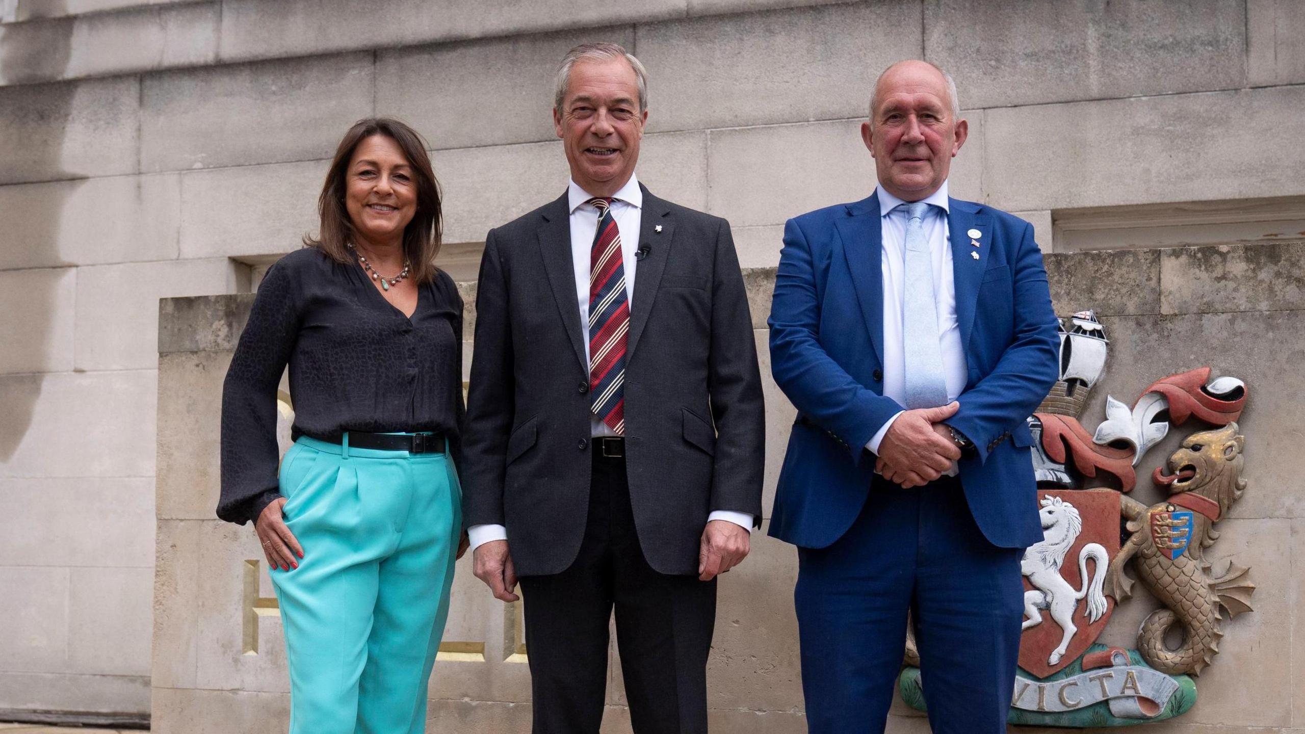 Linden Kemkaran wearing teal trousers and a black top. She is stood beside Nigel Farage, who has a red and navy tie and dark suit, and Brian Collins, who is wearing a dark blue suit. They are smiling in front of a stone building and a sign bearing the Kent coat of arms, which features a white prancing horse.