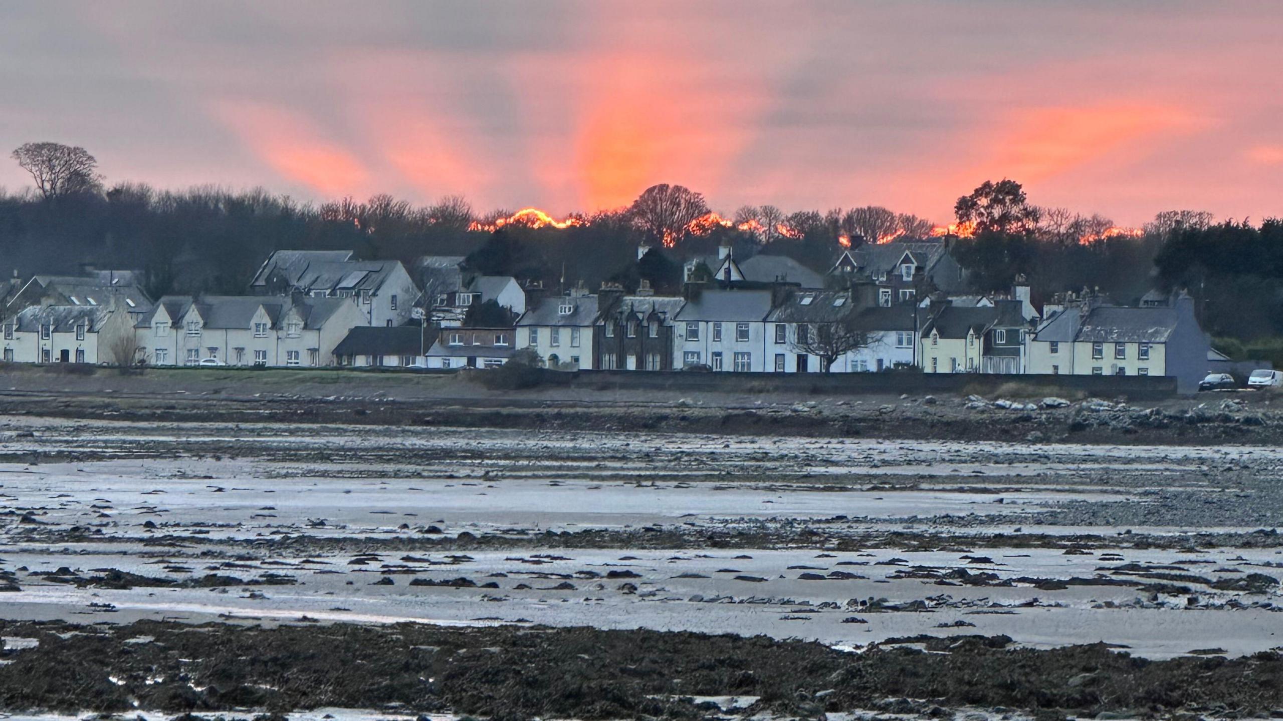 Orange rays over houses in Wigtownshire. The rays are orange and on the background of a lilac-grey sky. The houses in the foreground are all different colours including pale yellow, brown and white.