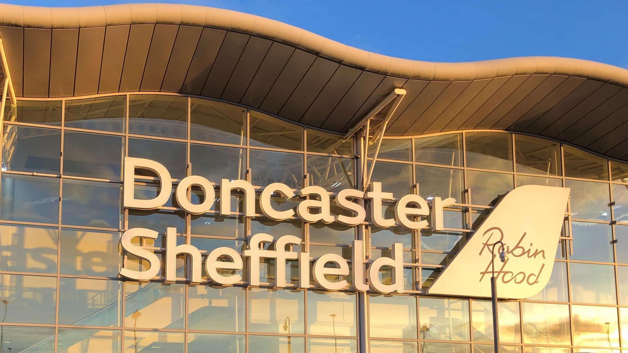 There are large white letters spelling out the words Doncaster Sheffield on a wall made up of glass. There is a logo in the shape of a plane's tail with the words Robin Hood 