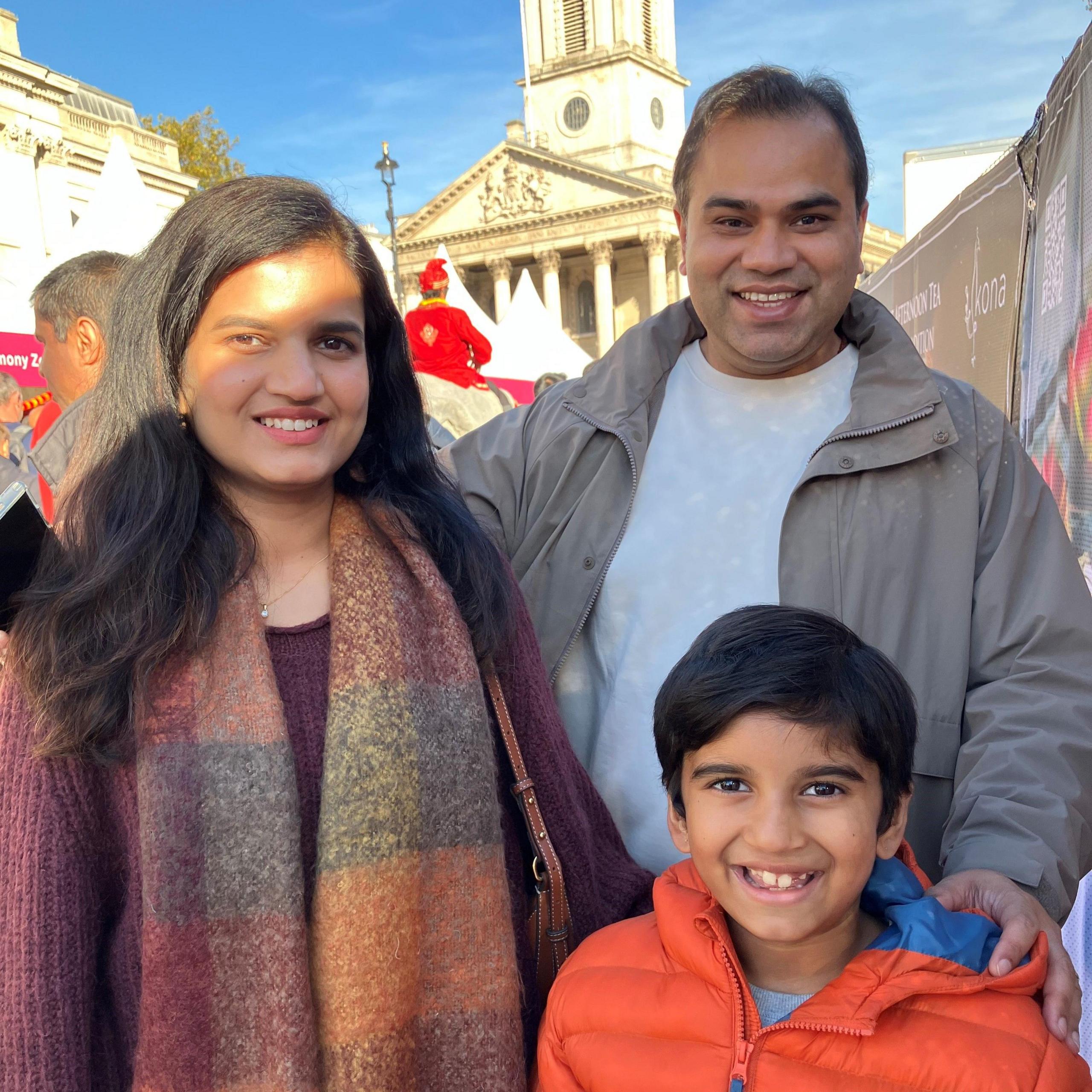 Ashu and Paritosh Gautam with their son Achintya, standing in front of the crowds in Trafalgar Square, with the steeple of St Martin-in-the-Fields visible in the background