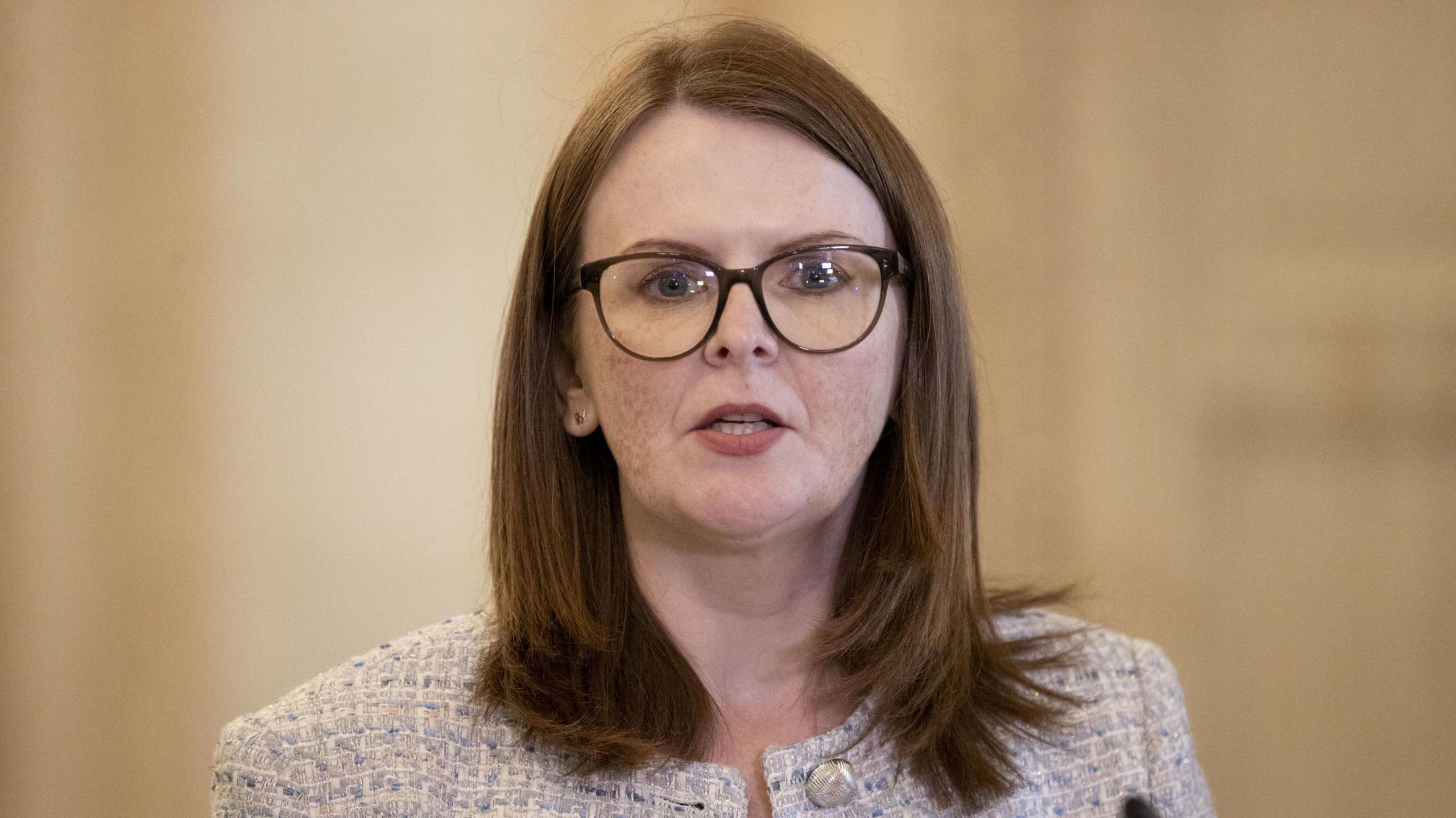 A woman with dark hair is wearing glasses and a grey jacket. She is in mid speech, and behind her is a beige wall. 
