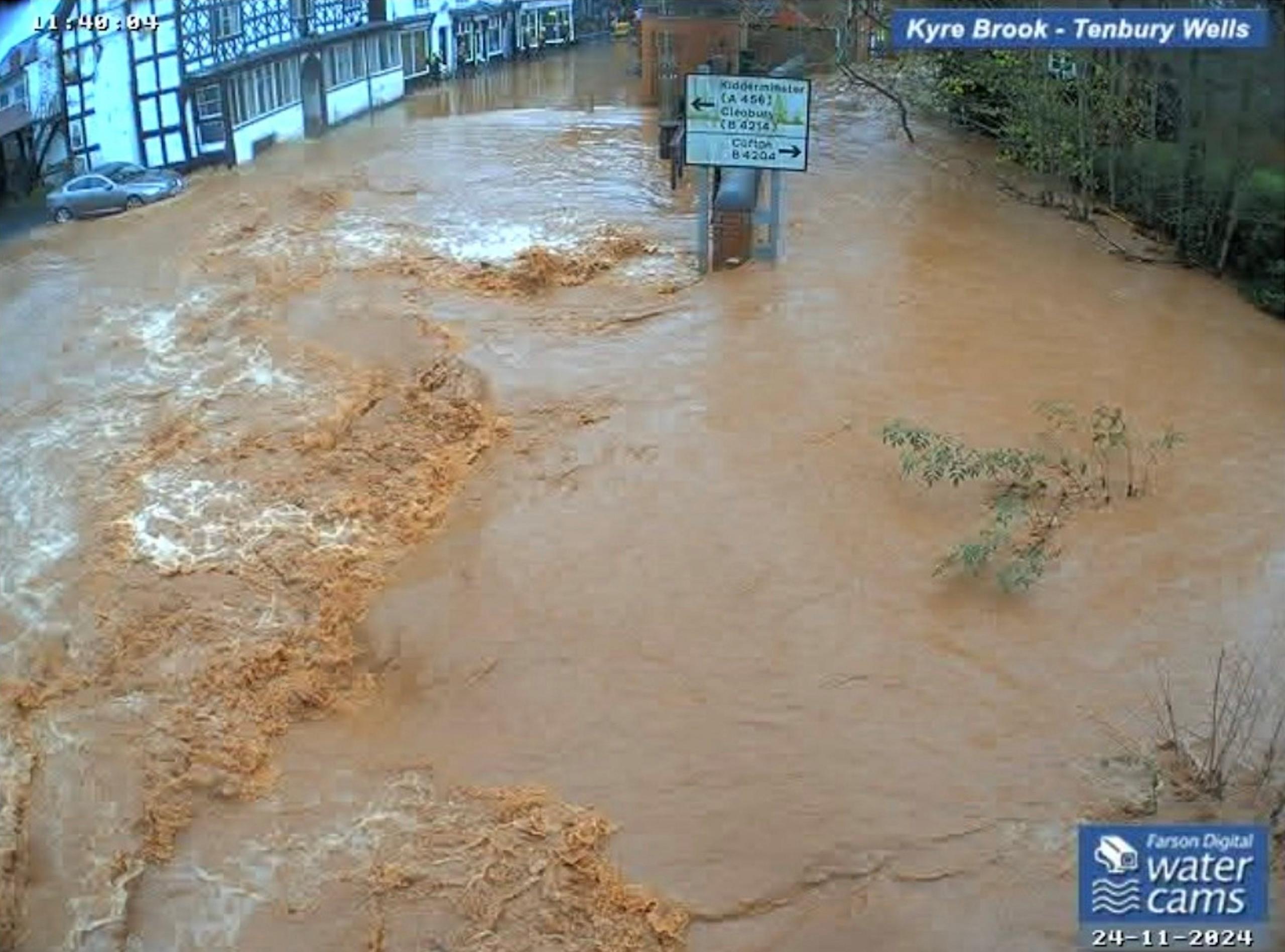 Brown-coloured flood water on a street after the wall collapsed. The legs of a road sign are partially submerged. A car is parked in the background near the water, next to a black and white building.