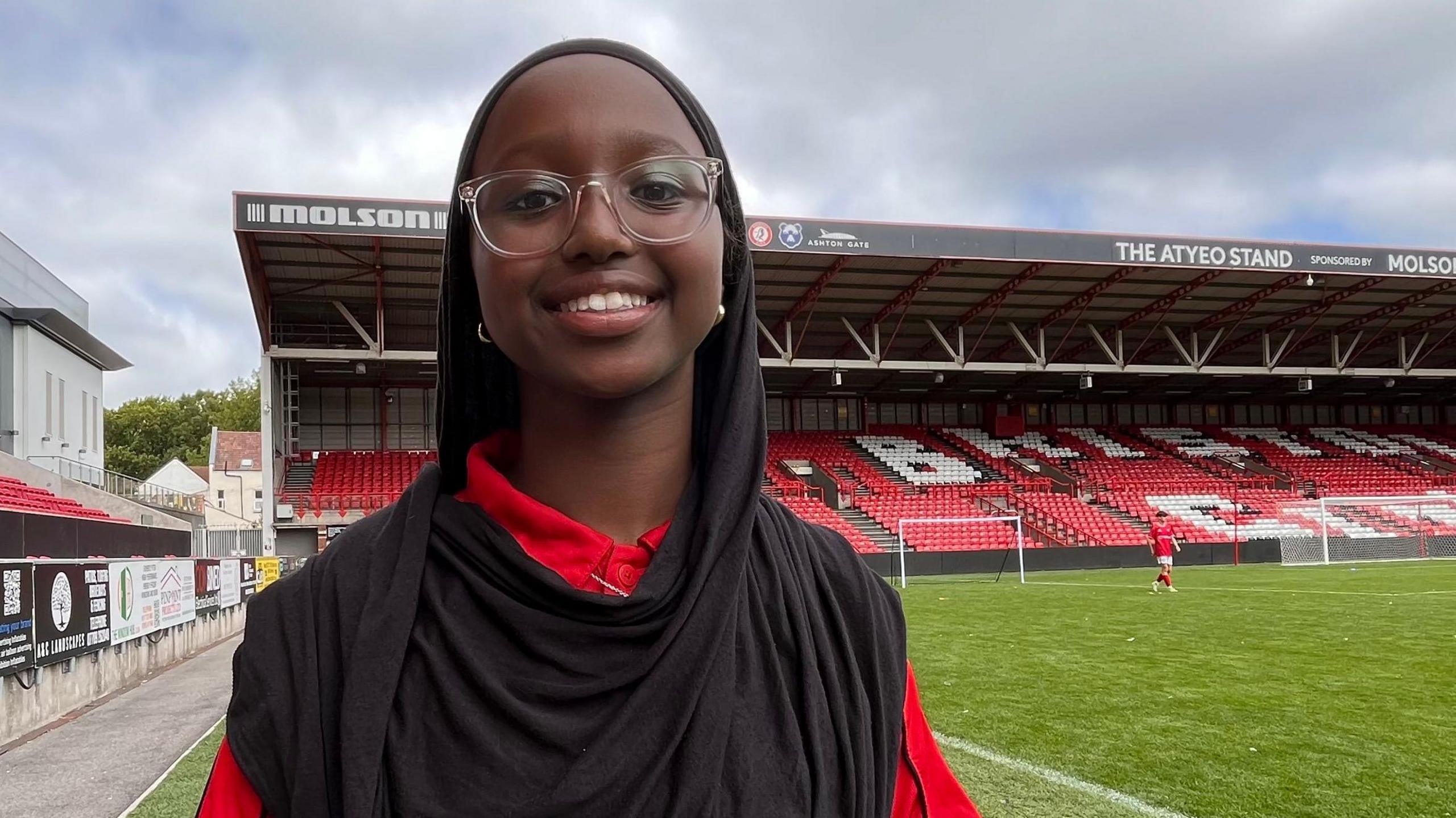 Mariam wears a red City Academy polo shirt, clear glasses and a black hijab. She smiles at the camera from the football pitch, which can be seen behind her.