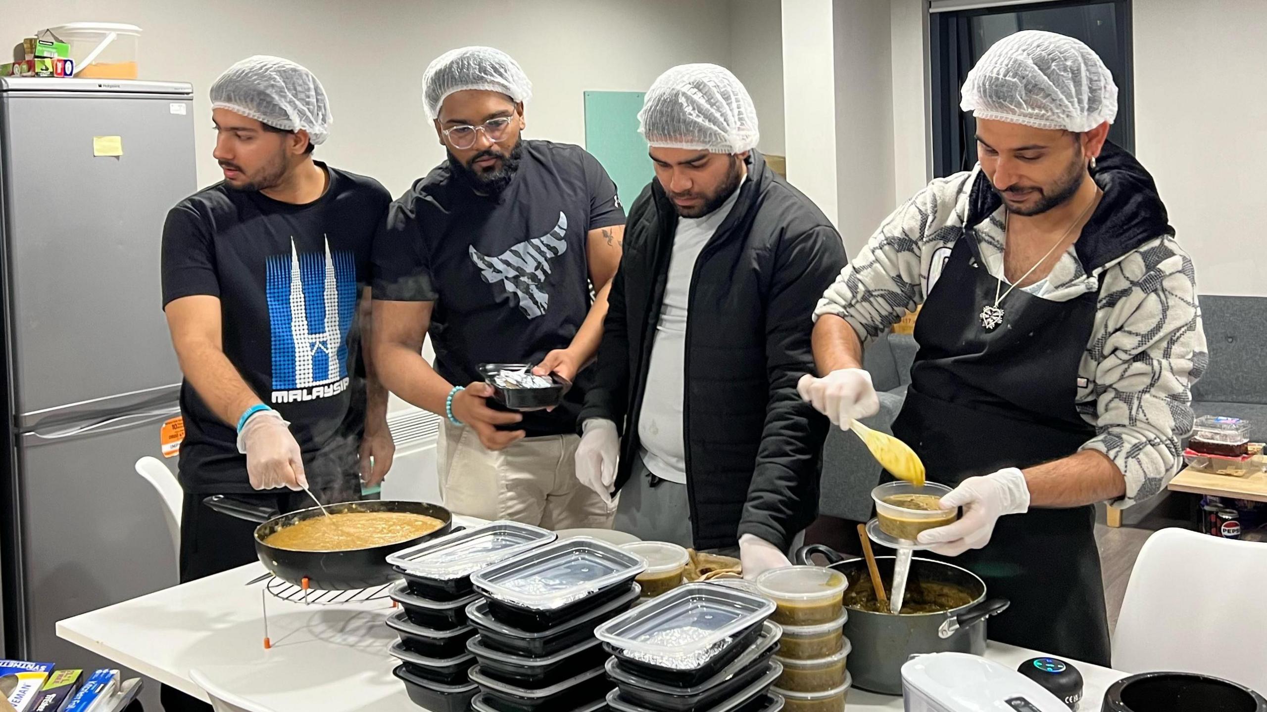 Four individuals with hairnets on preparing food. There are boxes for the food stacked on a table by two pots.