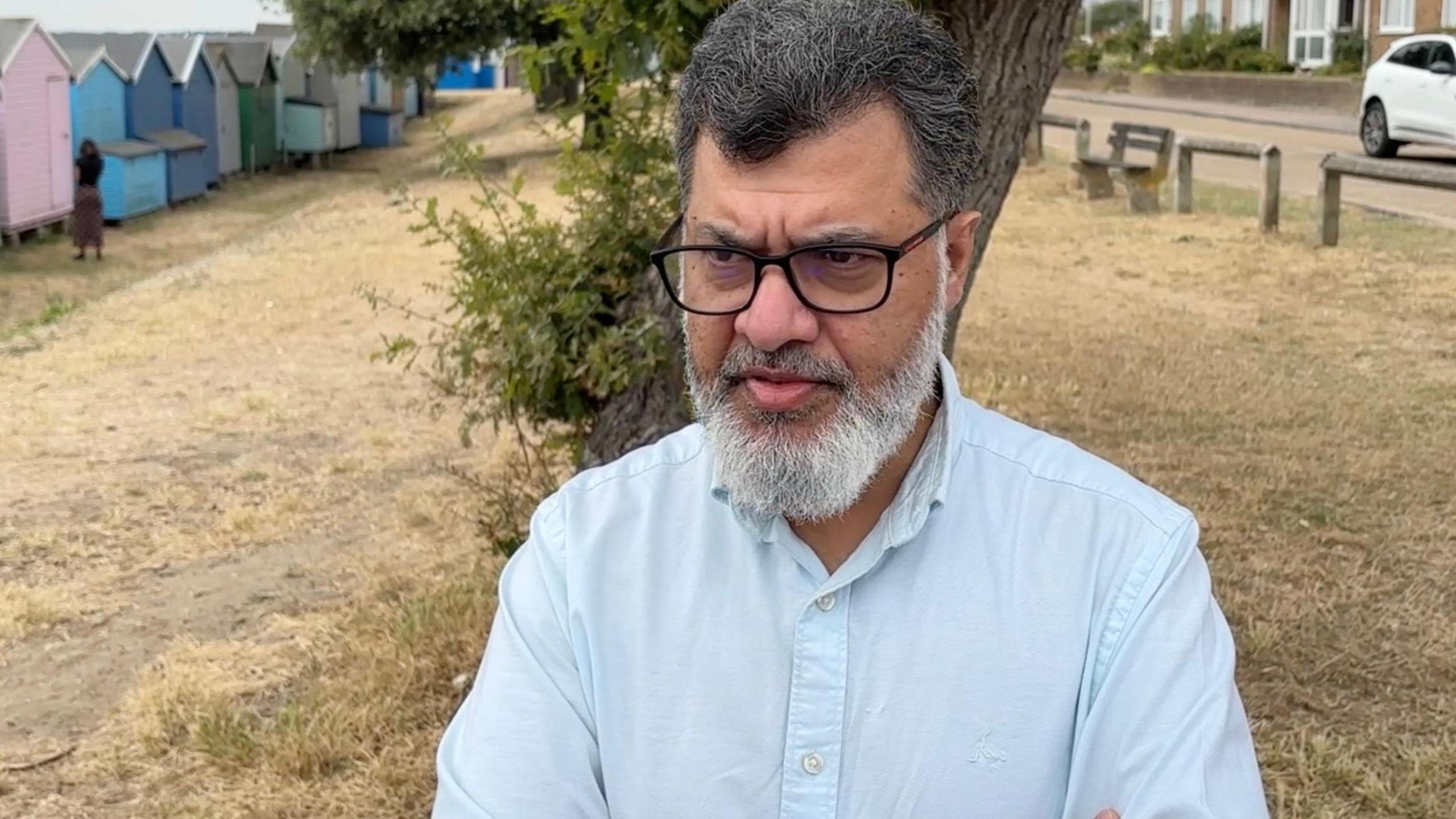 Mishba Uddin is looking away from the camera and wearing a blue shirt and has his arms crossed. He is wearing black framed glasses and has a beard.
He is standing on grass. There are beach huts to the left of him and there are homes on the right and a white car on the road.
