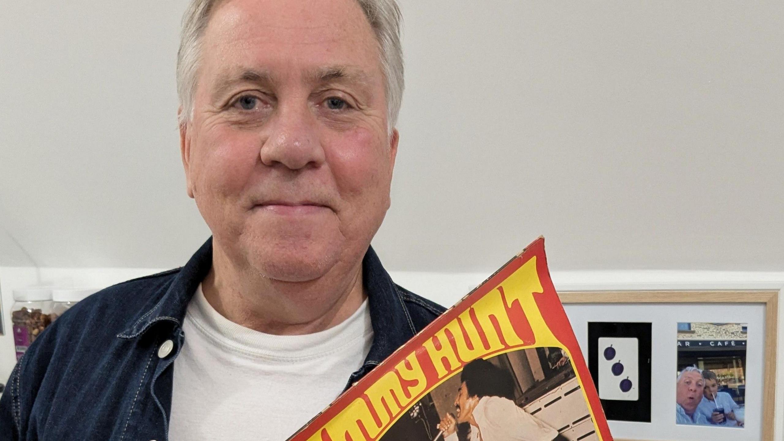 A man with grey hair, white T-shirt and denim shirt, holding a vinyl LP by American soul singer, Tommy Hunt