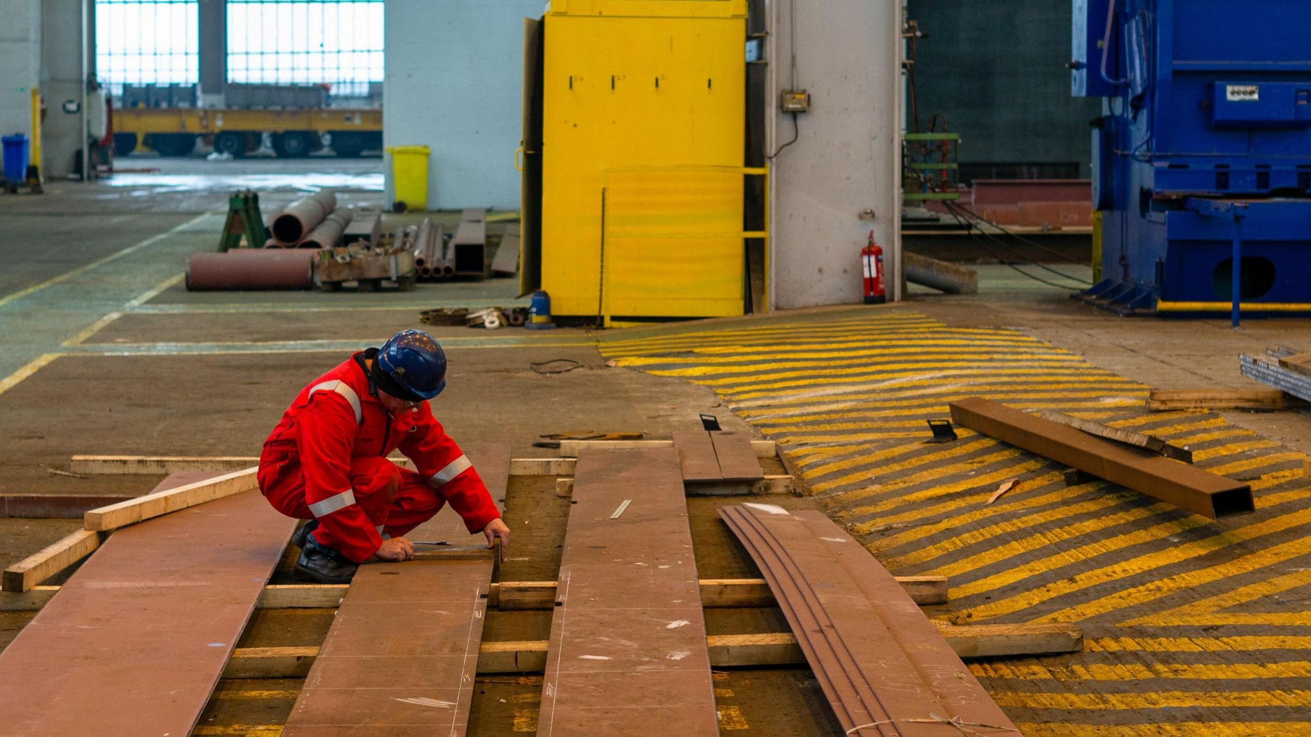 A worker measures pieces of steel for the construction of a barge at the production facility in Harland & Wolff Group Holdings Plc shipyard in Belfast. He's wearing a black hardhat and an orange high-viz clothes. There are pieces of tubes and wood lying about the floor.