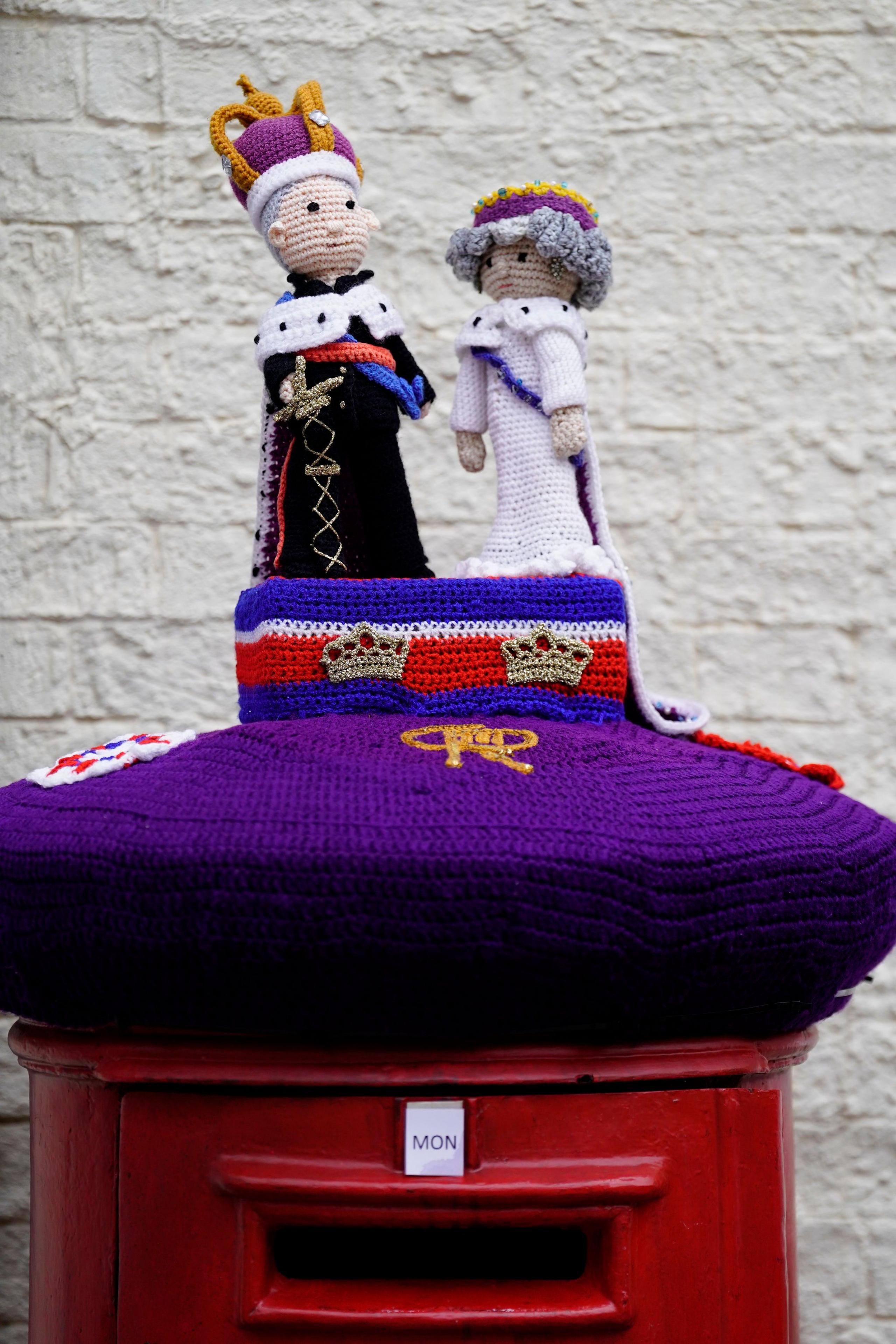 A woollen post box topper sees figures of King Charles and Queen Camilla standing on a plinth on top on top of a purple post box covering