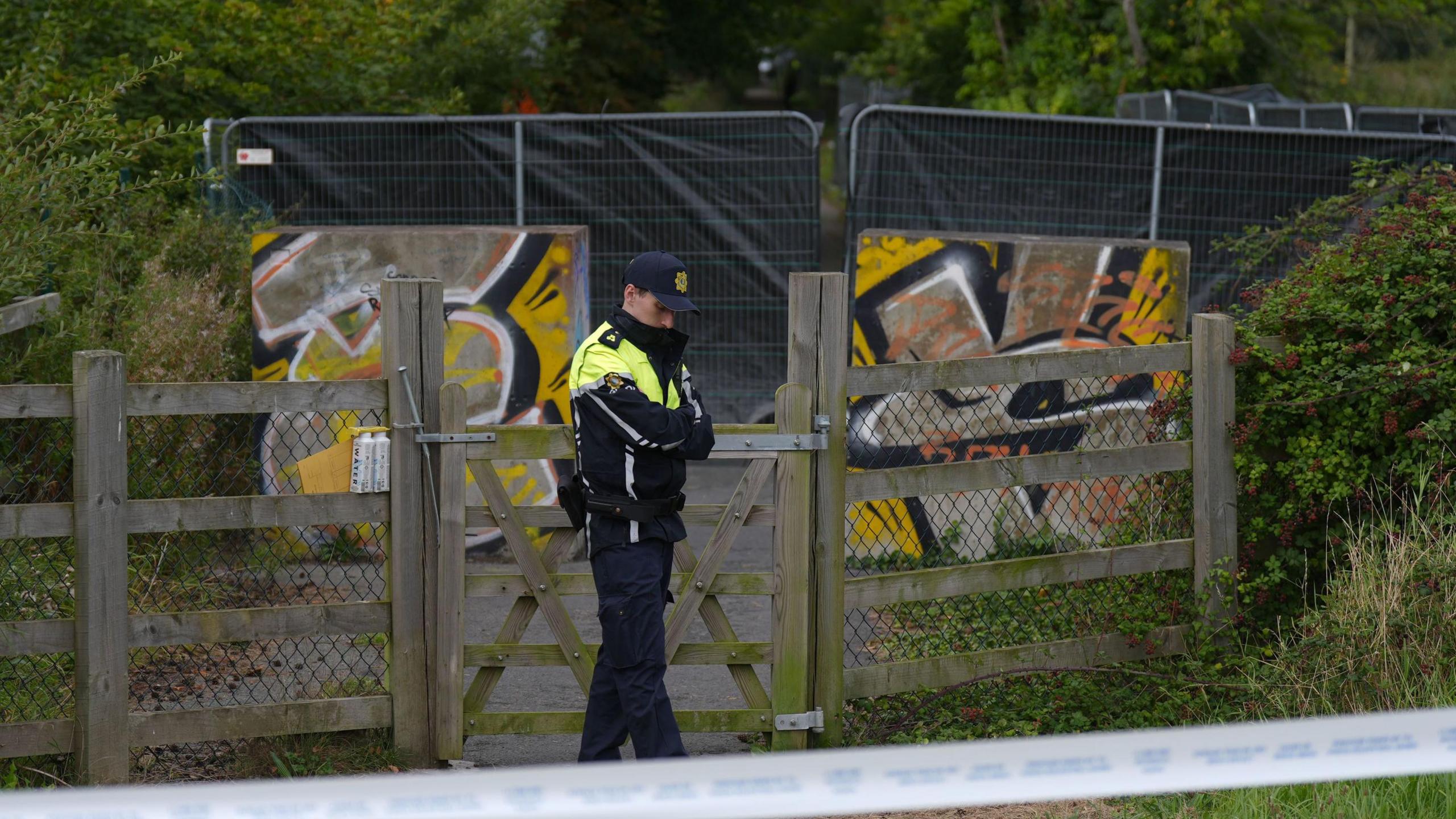 A man wears a black and hi-viz yellow jacket with black trousers. He wears a navy baseball cap. Behind him is a fence with a black sheet on it. protecting an area of green woodland.