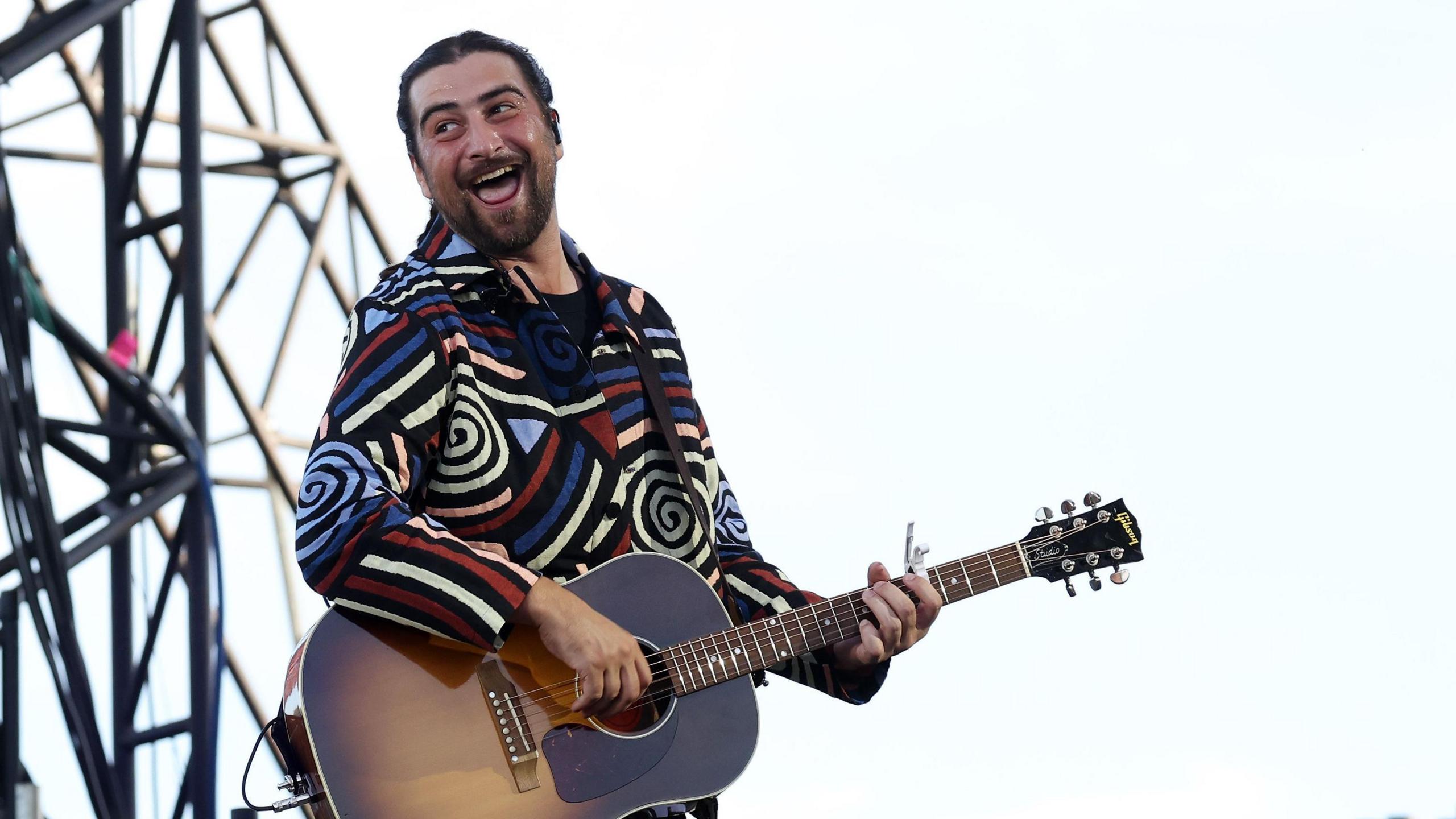 A man looks over his shoulder smiling as he sings. He is playing an acoustic guitar. He's wearing a bright, long-sleeved top with swirling colourful patterns on it.