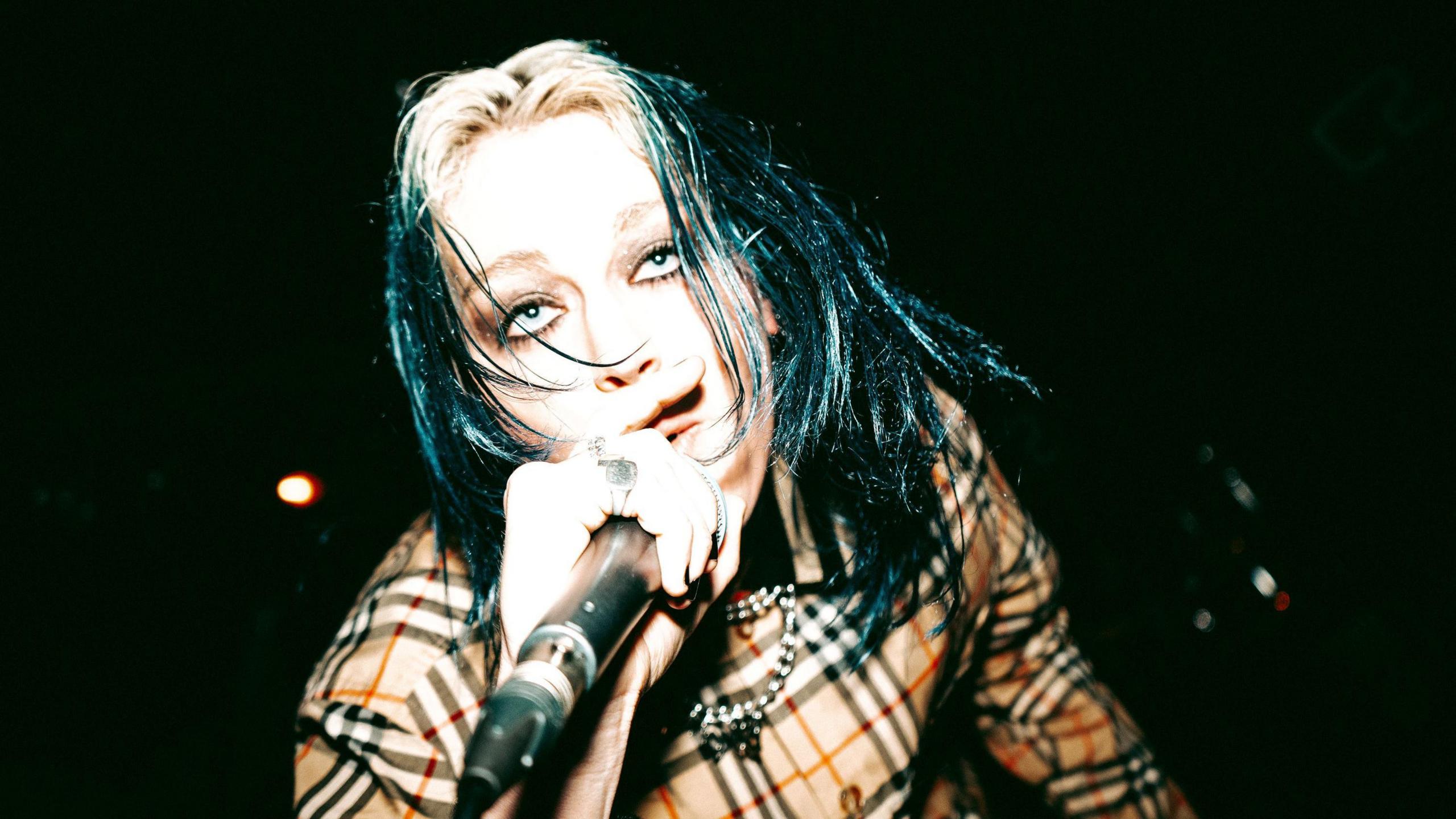 lleo wearing a Burberry shirt singing into a microphone. She has blue and blonde hair and dark eyeliner