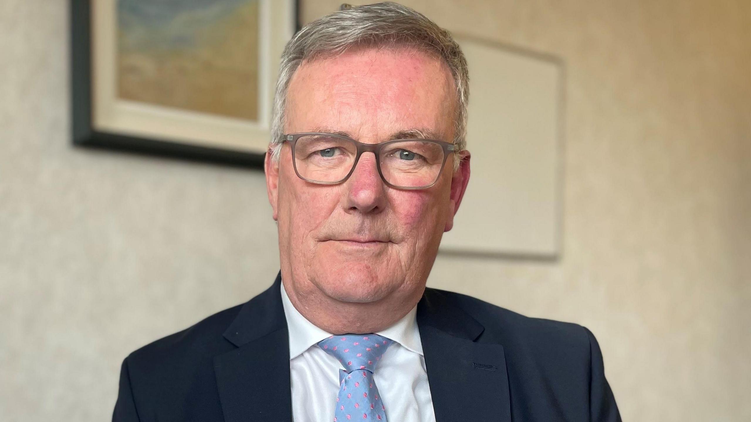 A man wearing a navy blue suit, white shirt and a pale blue tie. He is looking straight at the camera