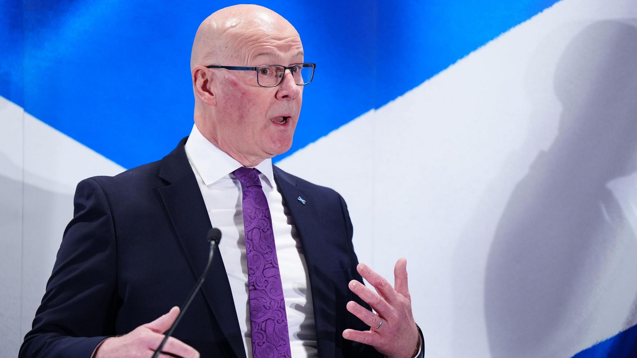 First Minister John Swinney speaking during a press conference at the Edinburgh Marriott Hotel Holyrood. He wears a suit with a purple tie and glasses.