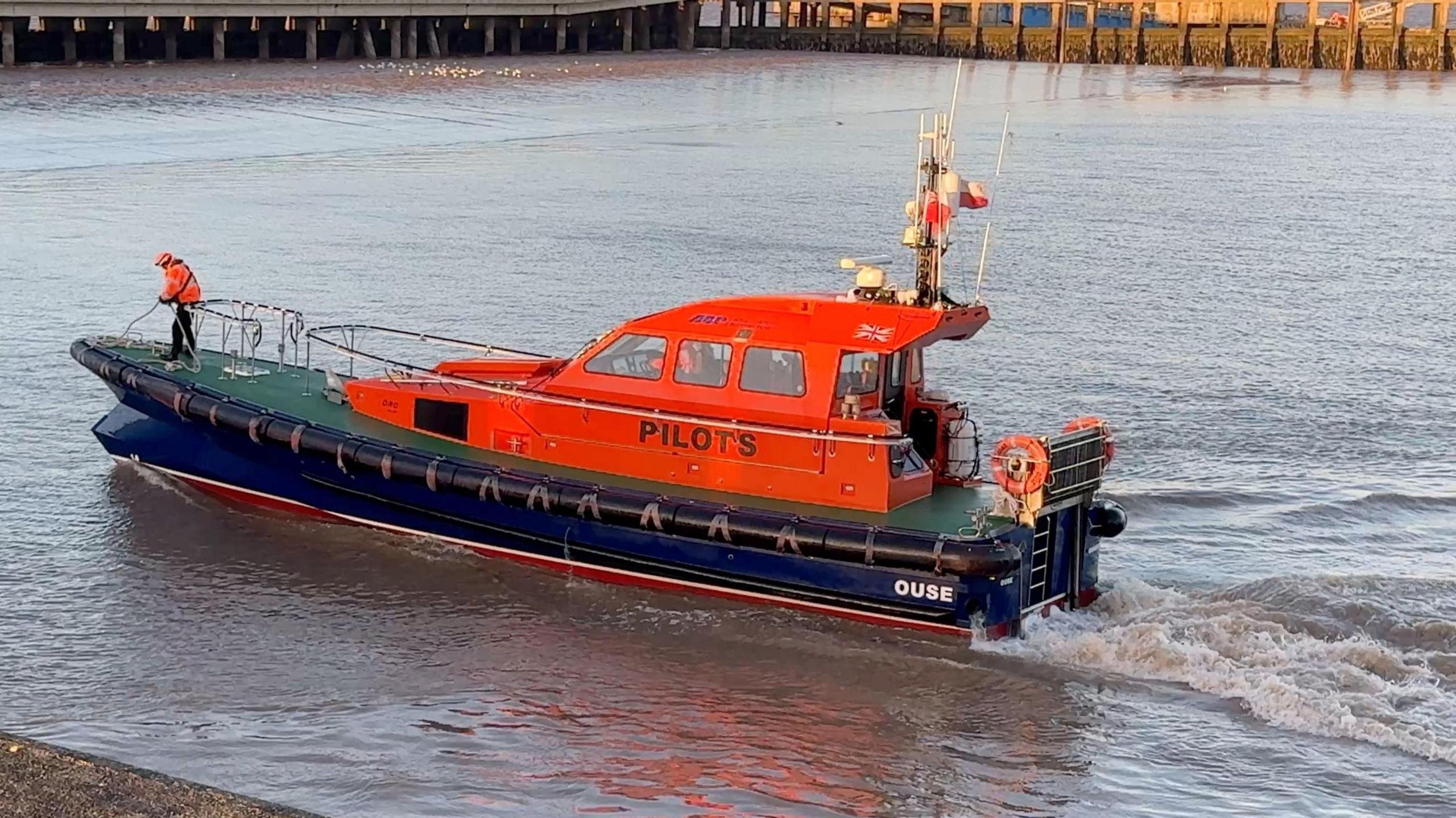 A man in an orange jacket stands at the prow of a fast launch ready to moor in Grimsby dock. The sleek launch has a blue hull and a dark orange-coloured cabin.