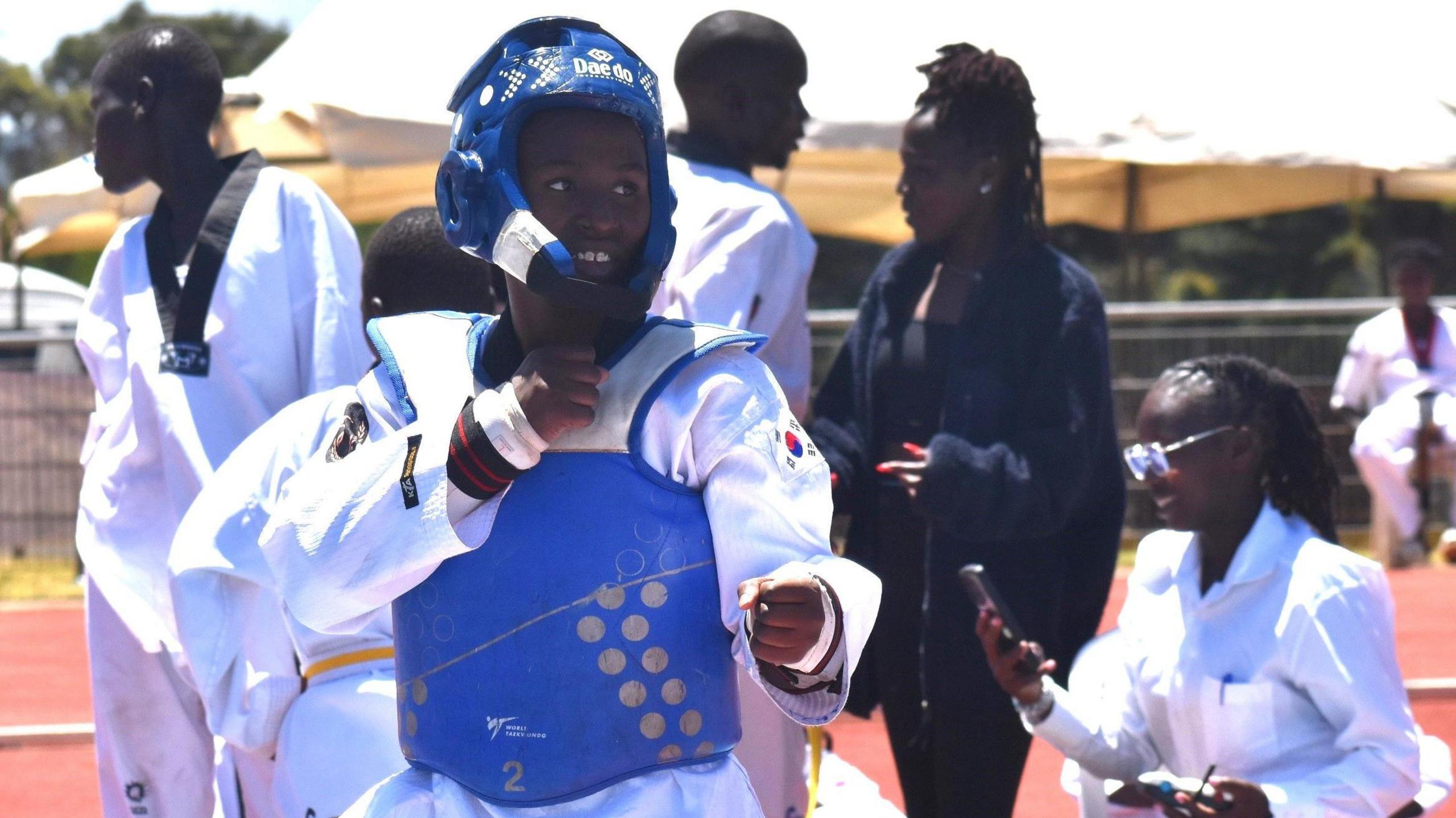 Laurence Namukiza wears a white taekwondo outfit and a protective blue helmet and chest guard as she takes a defensive pose while several competitors and officials are seen out of focus behind her