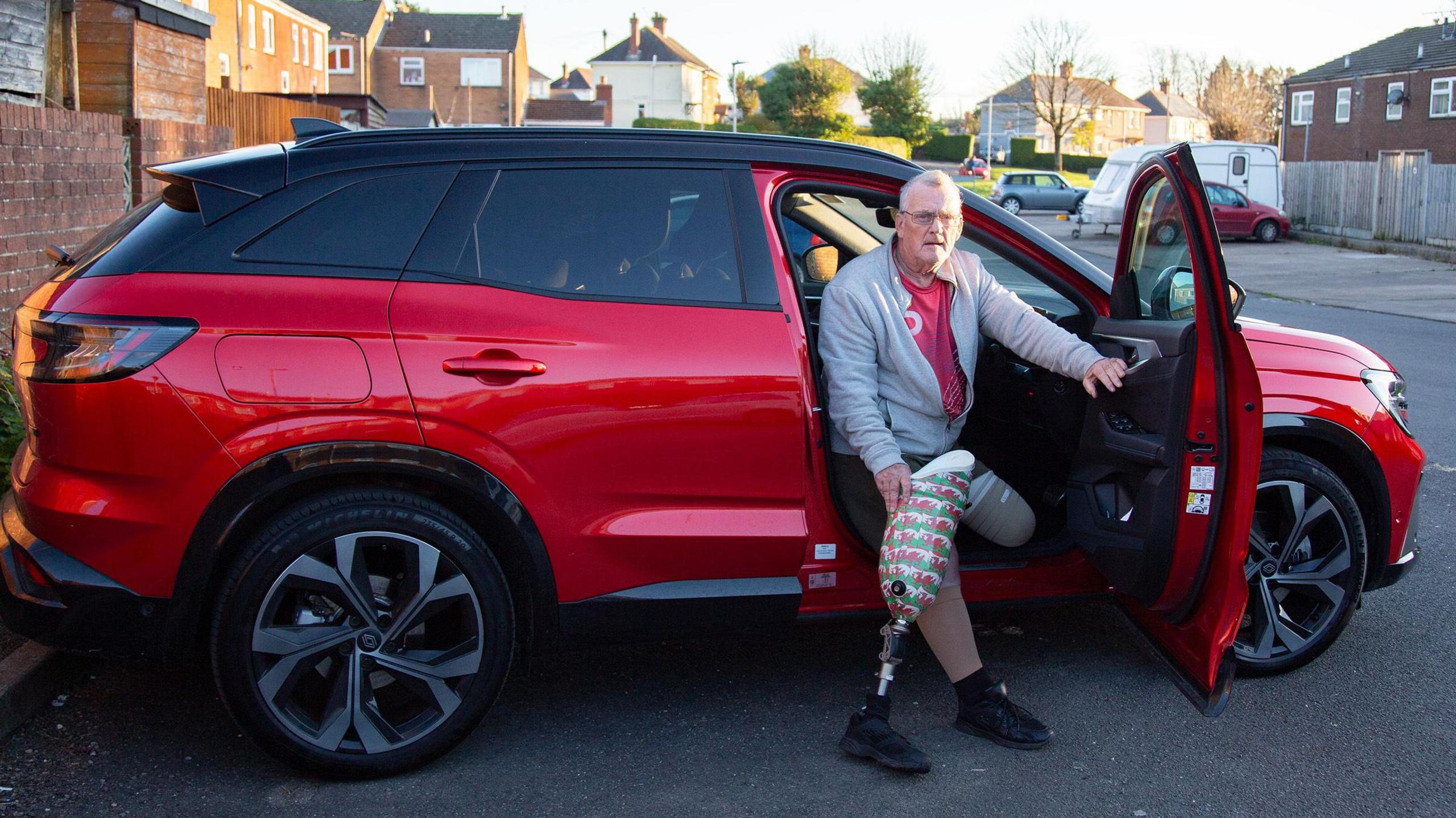 A man sits in the driver's seat of a large red car with the door open. The man is holding a prosthetic leg, and is wearing a grey zipped hoodie and shorts. 