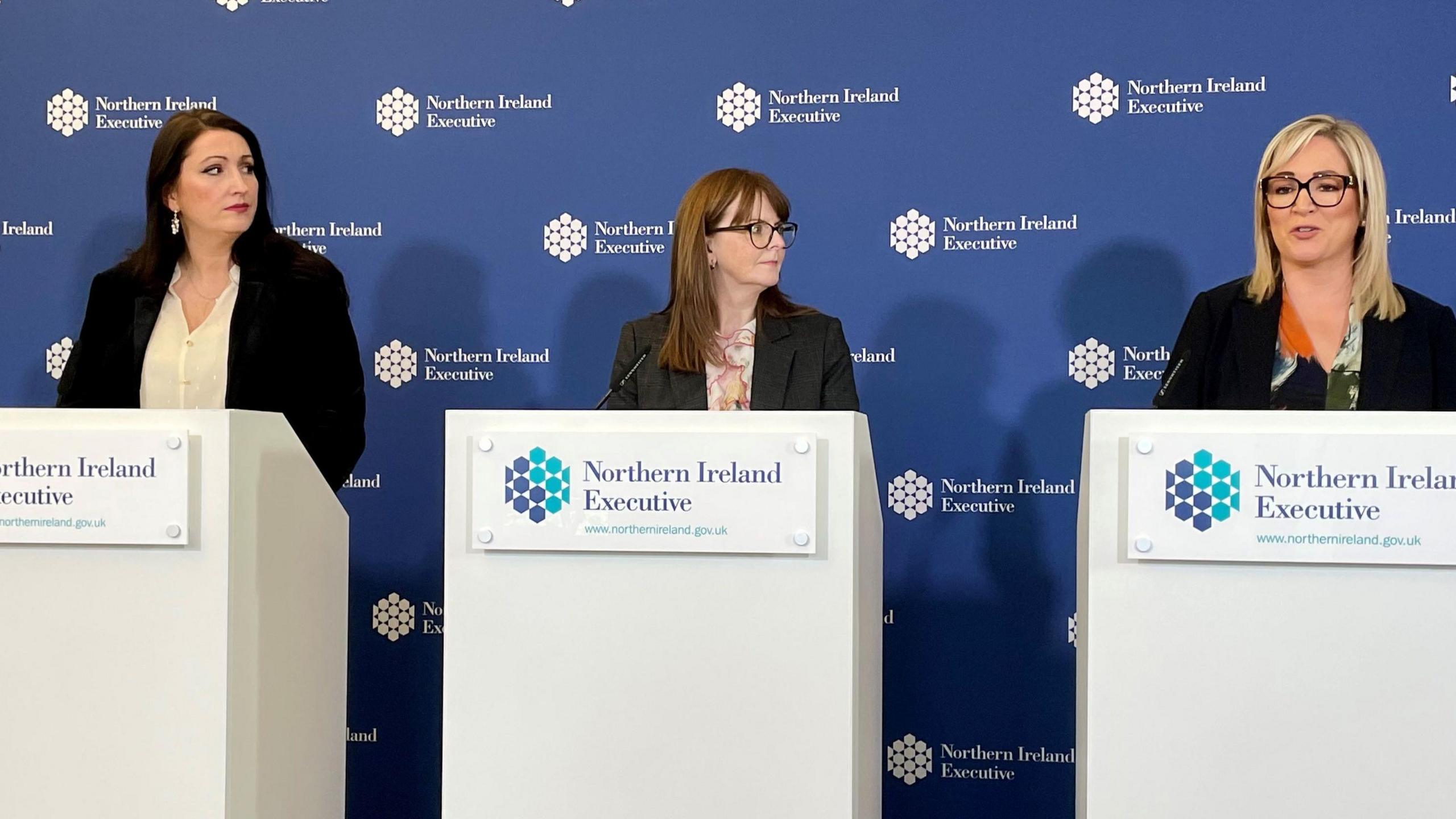 Deputy First Minister Emma Little-Pengelly, Finance Minister Caoimhe Archibald and First Minister Michelle O'Neill in black blazers, stood in front of Northern Ireland Executive white podiums with microphones, against a blue backdrop