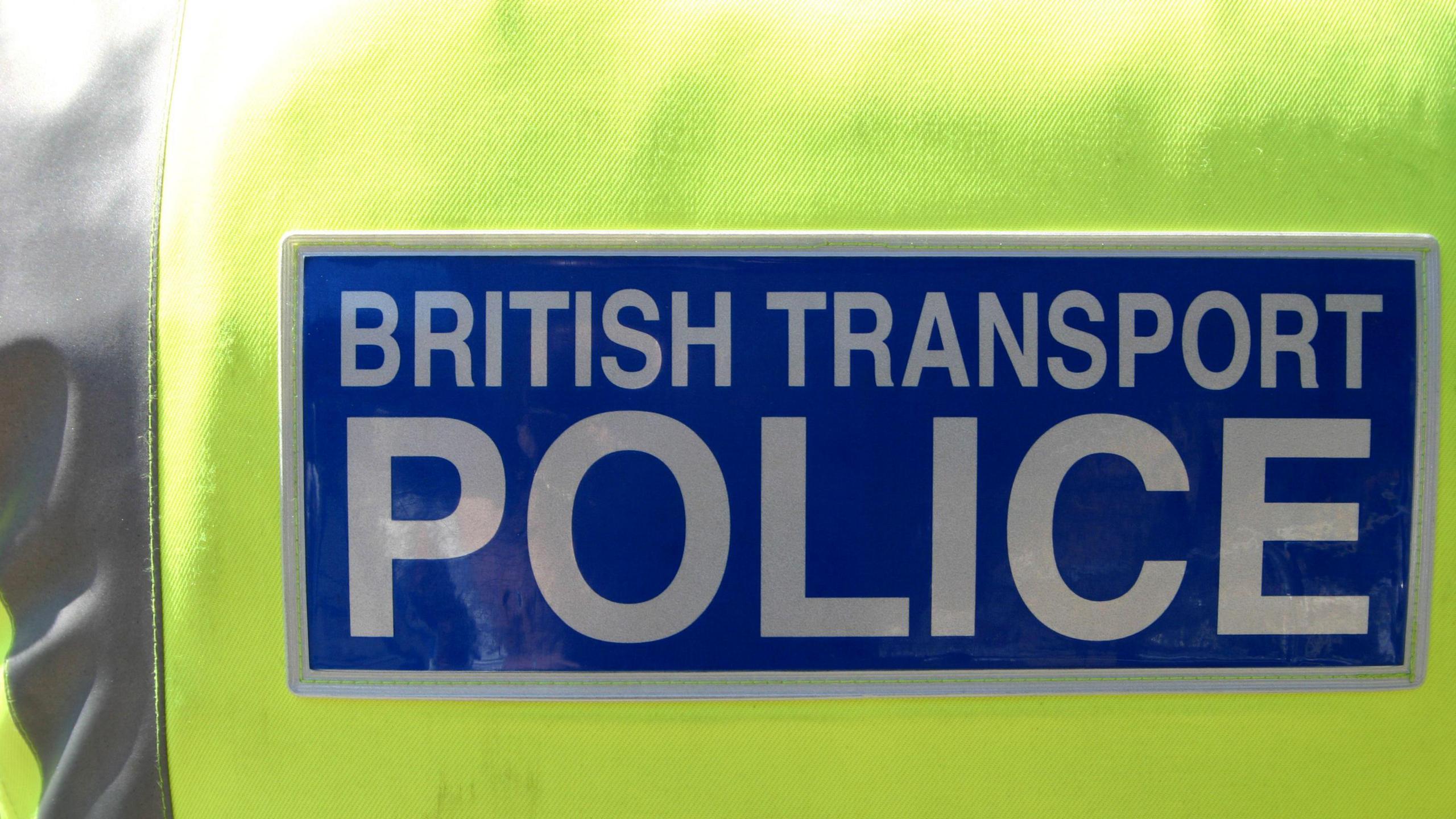The back of an officer's yellow hi-vis. In a blue box, it has the words 'British Transport Police' in white bold letters. To the left is a silver strip down the jacket.