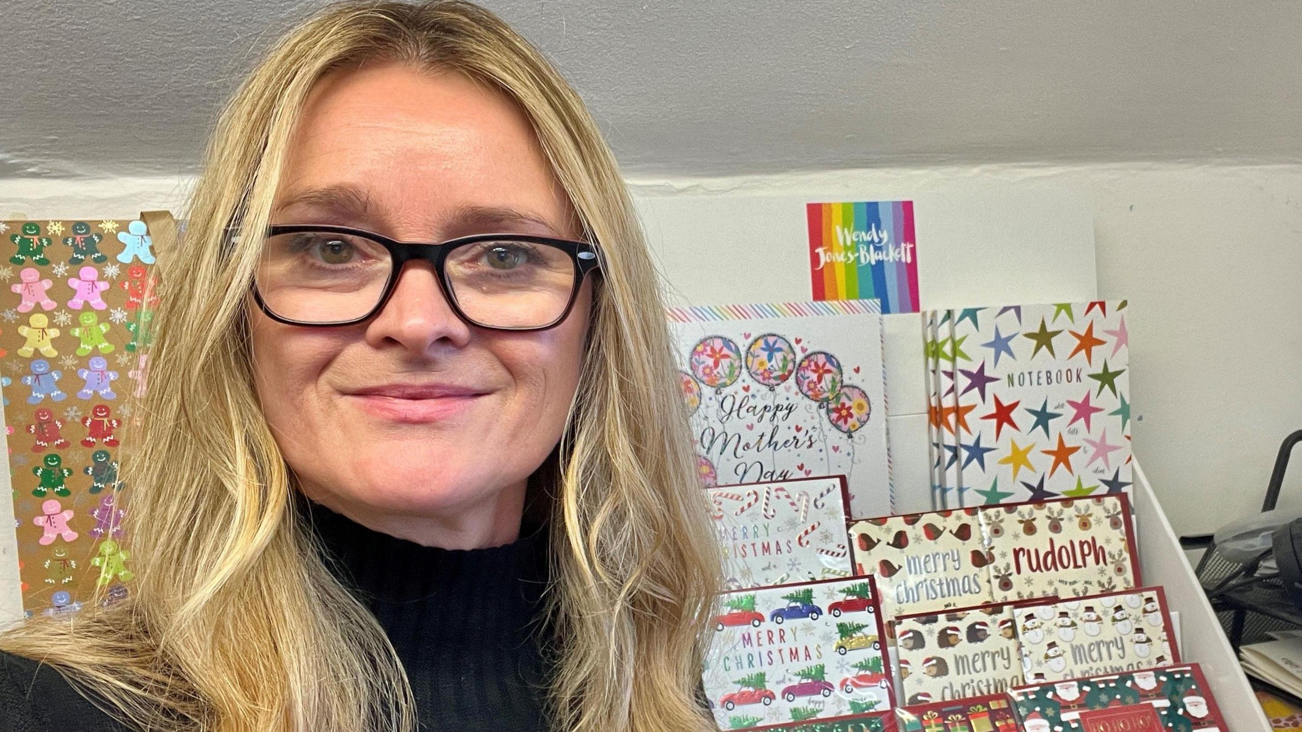 Wendy Jones-Blackett looks down the barrel of the camera as she poses in front of a selection of greeting cards covered in stars, cars and Christmas trees. She is wearing a black turtleneck jumper and has loose blonde hair and black-framed glasses