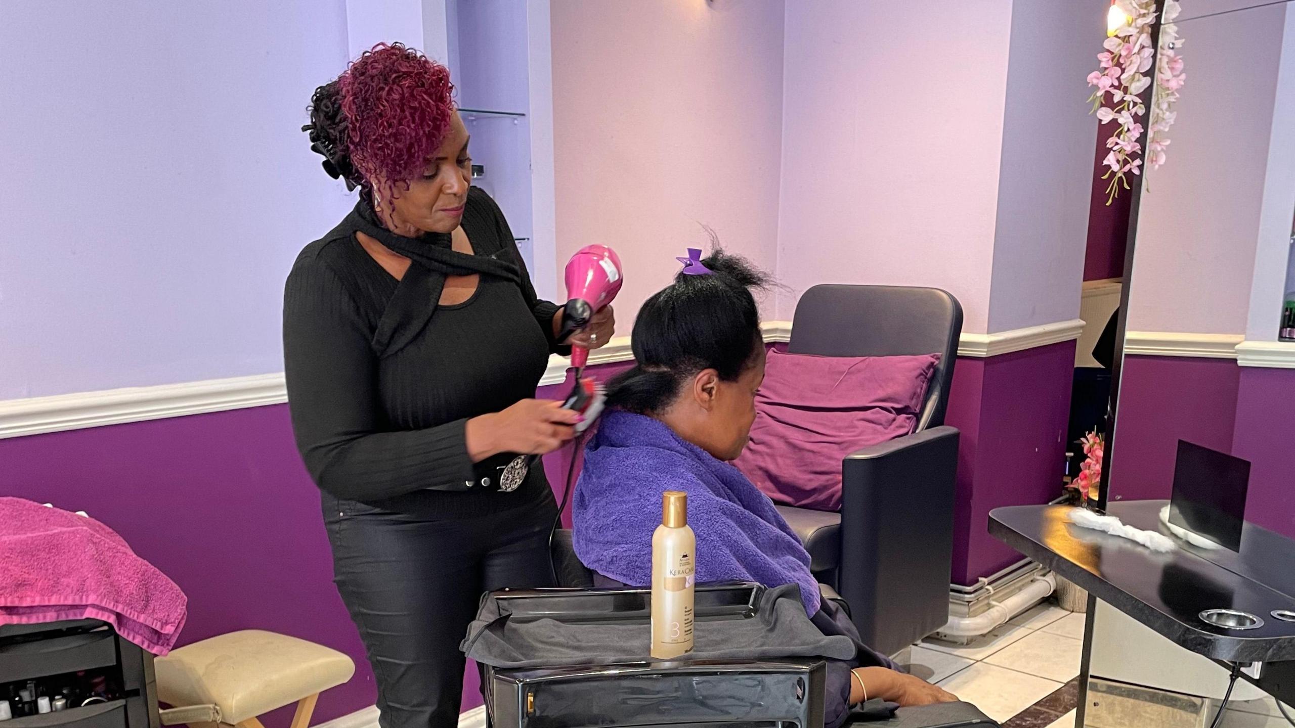 Wilma Burke styles client Katherine Kidd’s hair using a pink handheld hairdryer. Katherine sits in a black salon chair at the centre of the salon, while Wilma stands behind her. The walls and décor feature purple, burgundy, and black tones.
