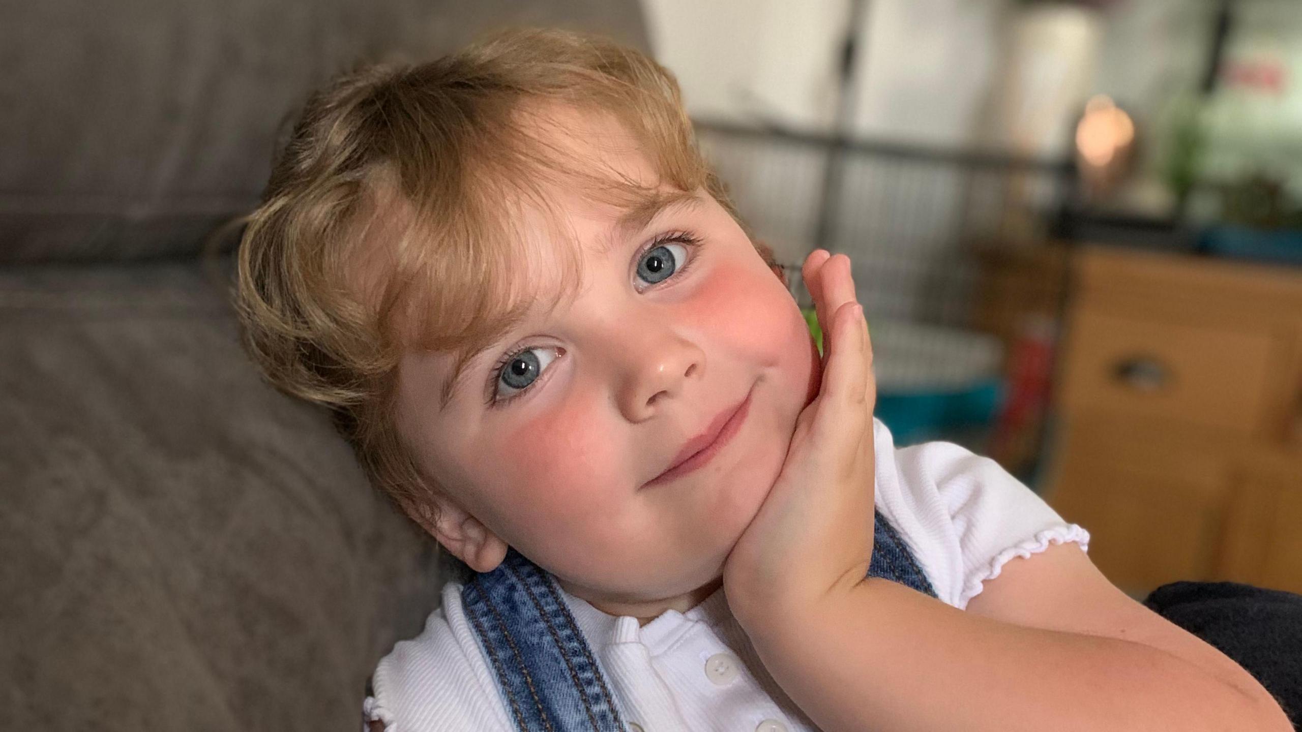 Amber is smiling at the camera as she leans her head back on a sofa and rests her chin on her left hand. She has clear blue eyes and long blonde hair tied back from her face. She is wearing a short-sleeved white T-shirt and there are two denim straps over a shoulder.