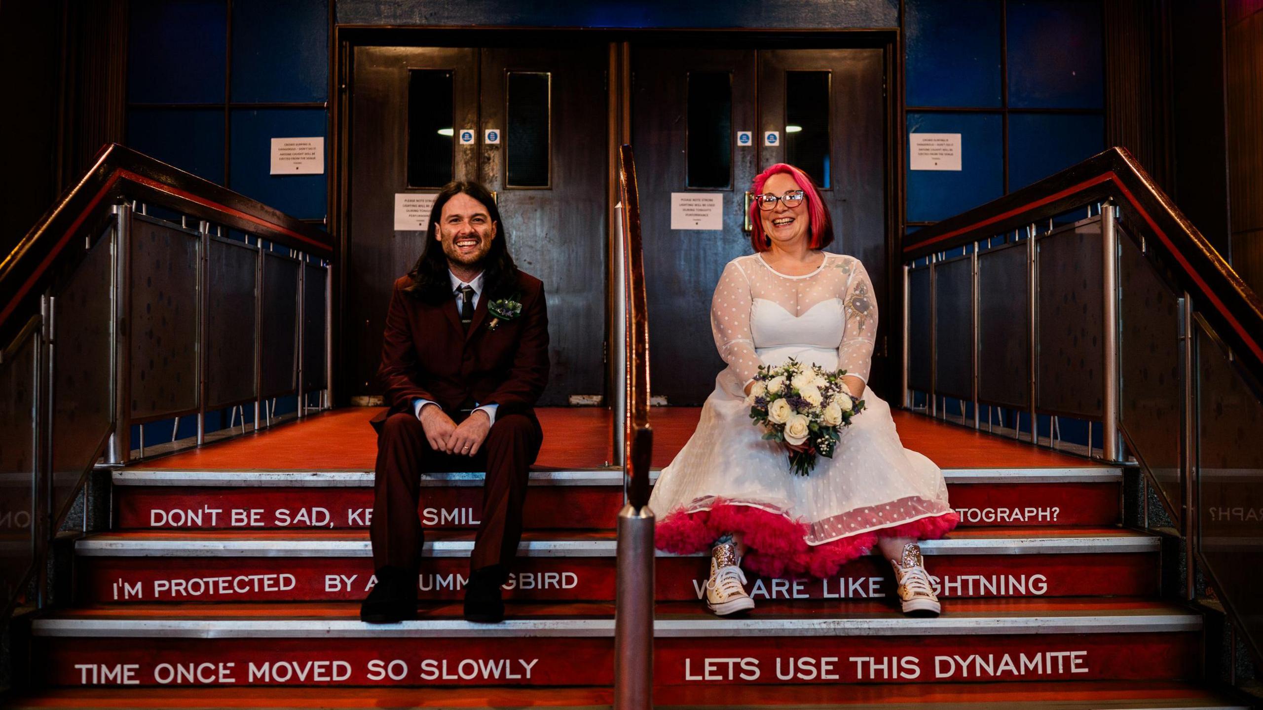 Bride and groom sitting on steps in the Barrowland Ballroom with song lyrics written on each step, both wearing their wedding attire