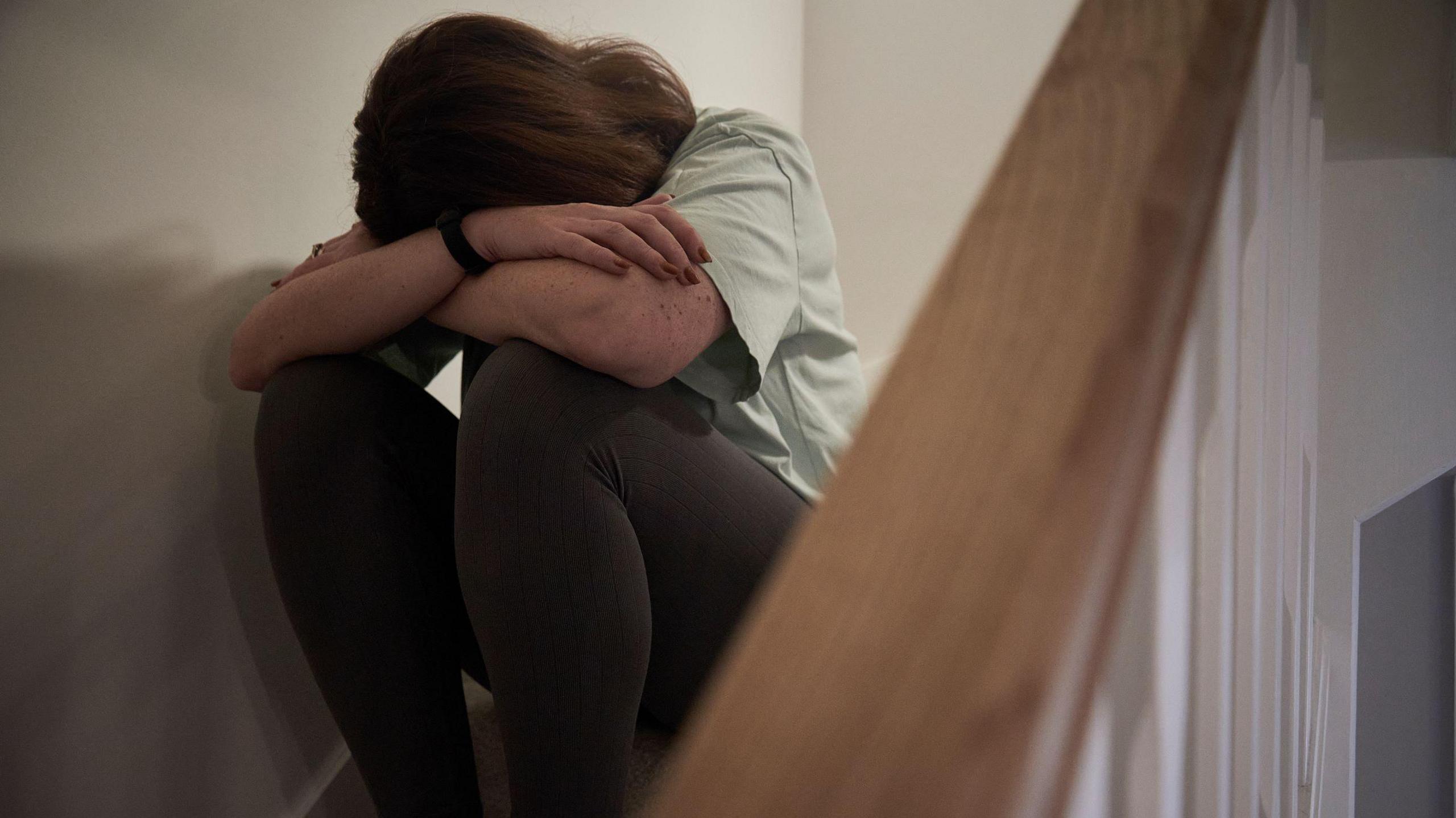 A woman sitting with her head in her hands on the stairs of her home. She has brown hair and is covering her face. 