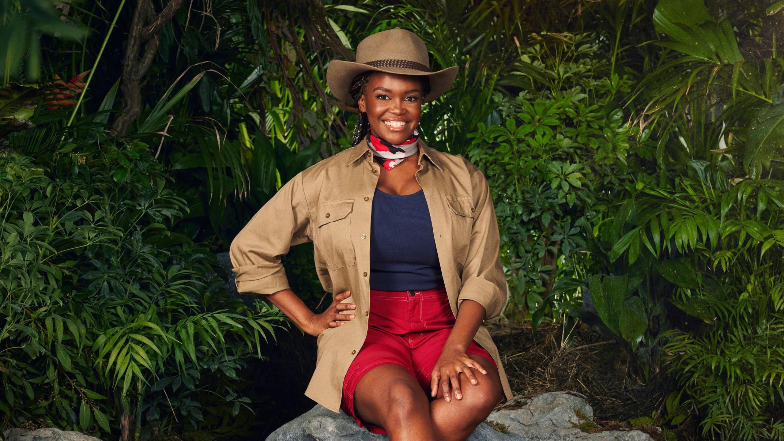 Oti Mabuse wearing her jungle outfit, sitting on a rock surrounded by plants