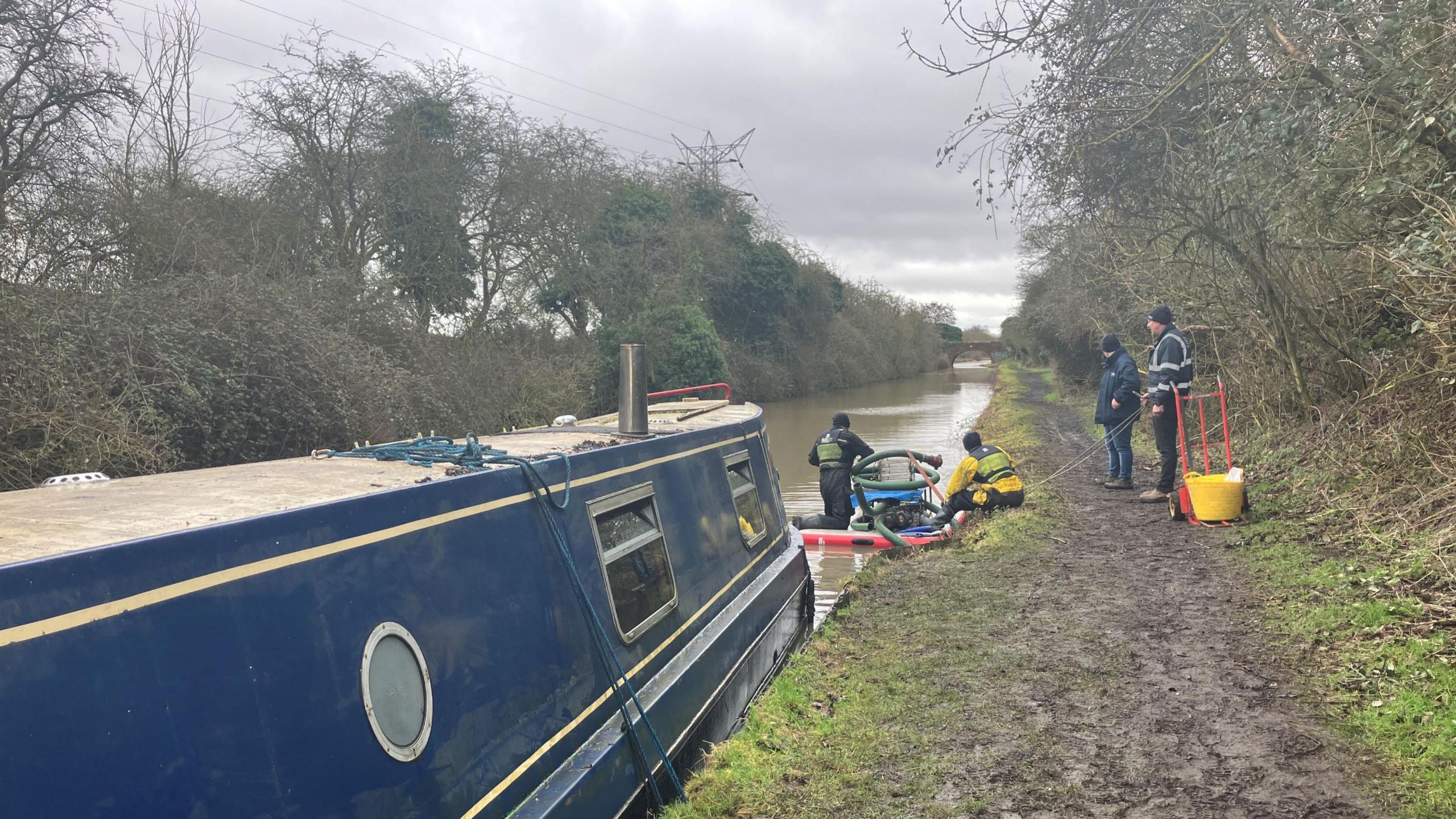 Two men on the right in coats and hats stand on the towpath by a blue narrow boat. Two more people are on a raft in the water in front of the boat.