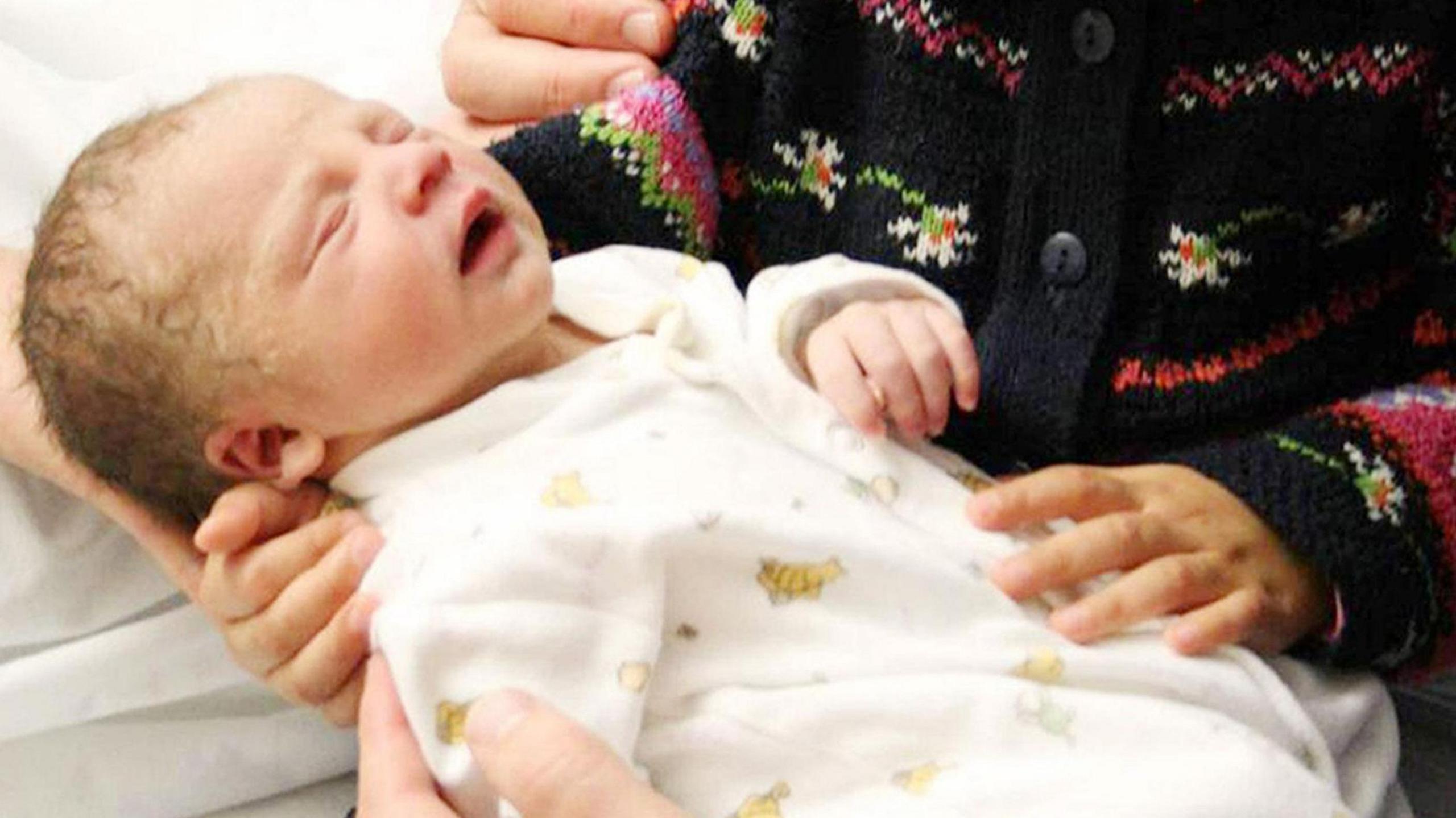 Joshua Titcombe before his death at just nine days old. He is wearing a white onesie with characters printed on it. He is being held by a child wearing a colourful knitted cardigan.