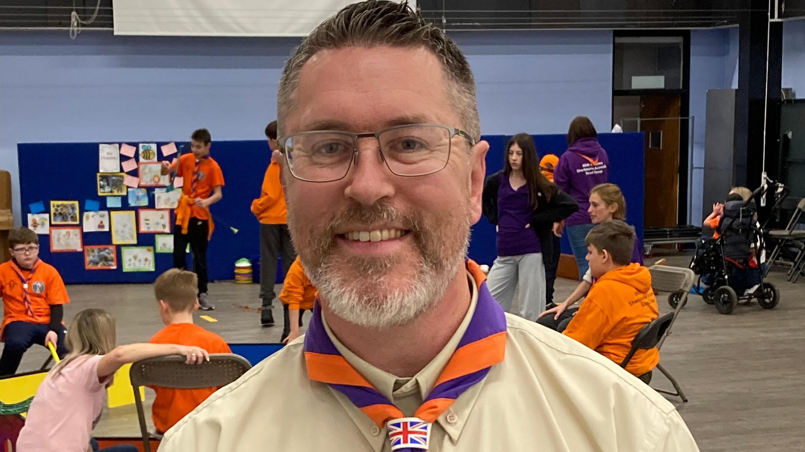 A man with short hair and a short, greying beard. He is wearing a light brown short with a purple and orange neckerchief. Behind him there are several children in orange hoodies taking part in activities.