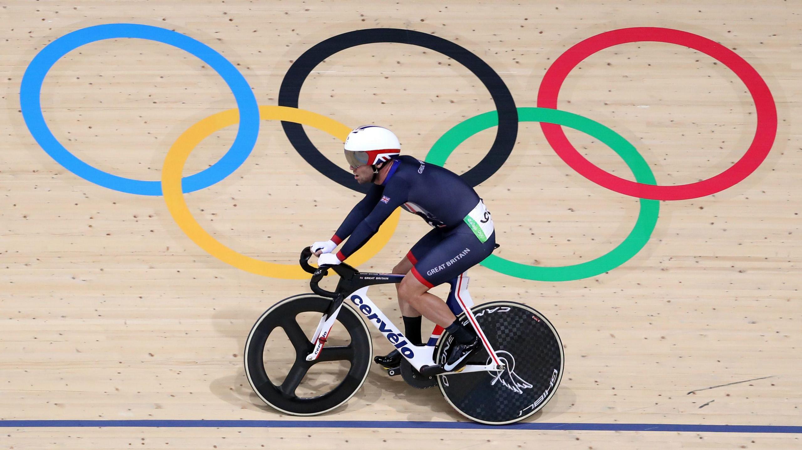 Sir Mark Cavendish, wearing a white helmet with a Union Jack on it and a blue cycling kit, cycles on a black and white bicycle on a wooden track with a blue line on it and the five Olympic rings