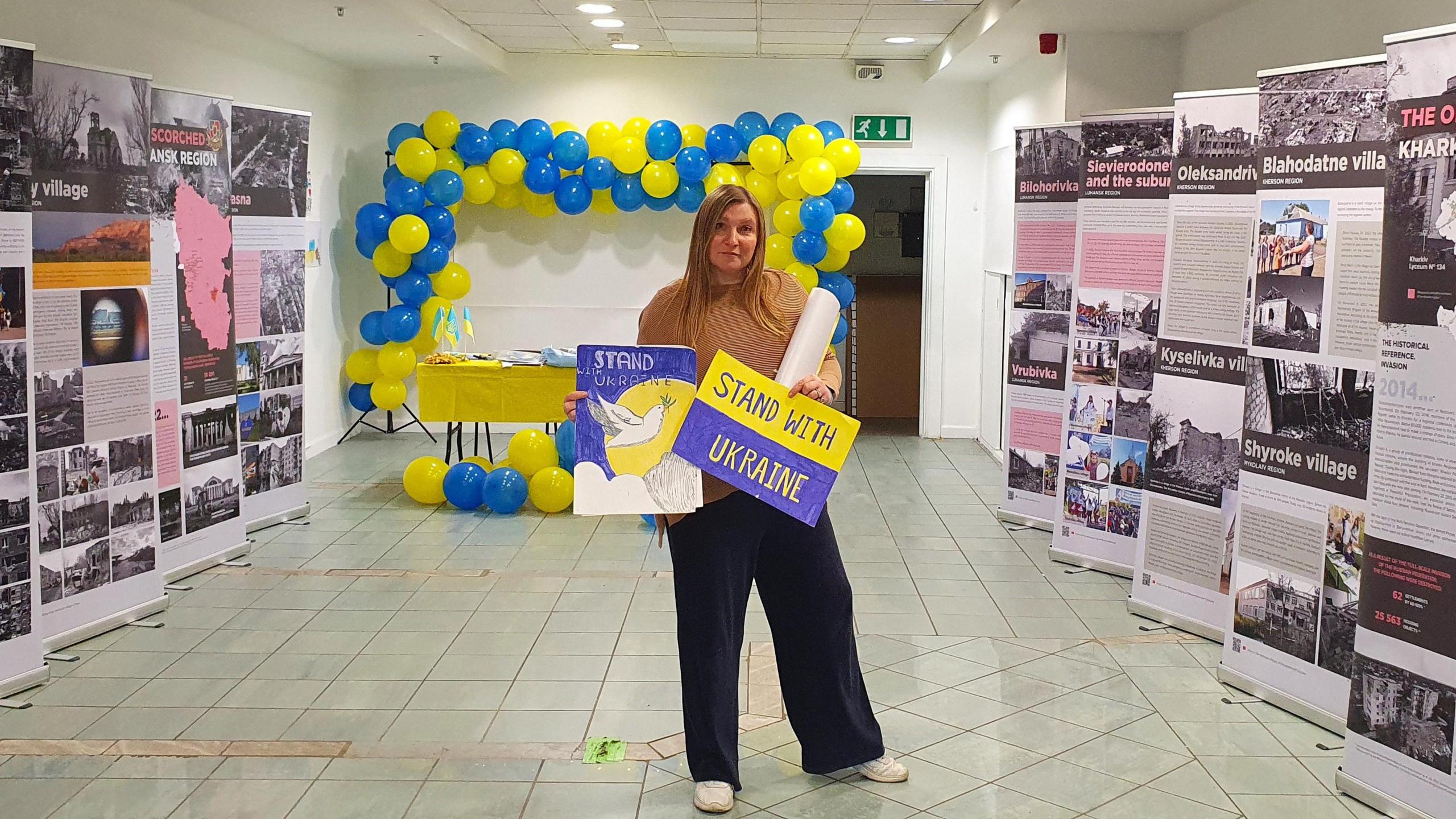 Antonina Grebeniuk wearing wide leg trousers and a brown jumper. She is standing inside a white room in Broadmead shopping centre, holding two hand painted posters of the Ukrainian flag colours that say 'Stand with Ukraine'. On either side of her there are banners full of images showing the destruction of Ukrainian towns, cities and villages. 