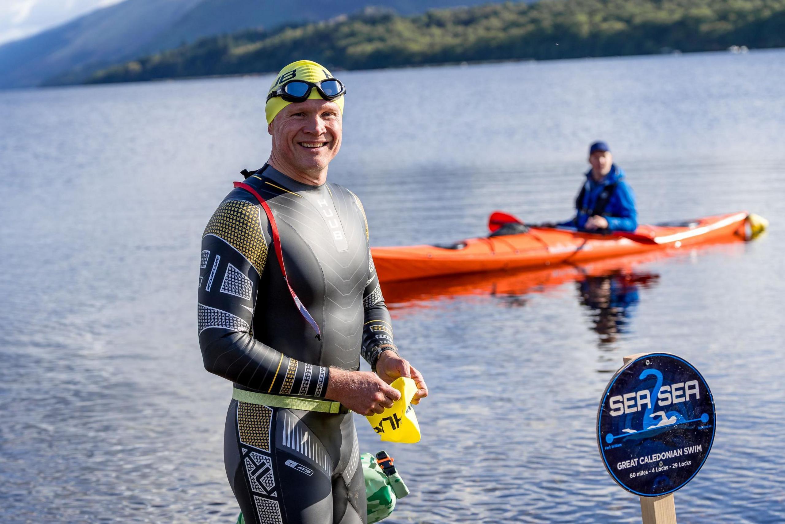 Iain is standing out of the water in a black wet suit, yellow swim cap and his goggles on his forehead. Behind him in the water is a member of his support team in an orange kayak. A round sign show the name of Iain's challenge: Sea2Sea.