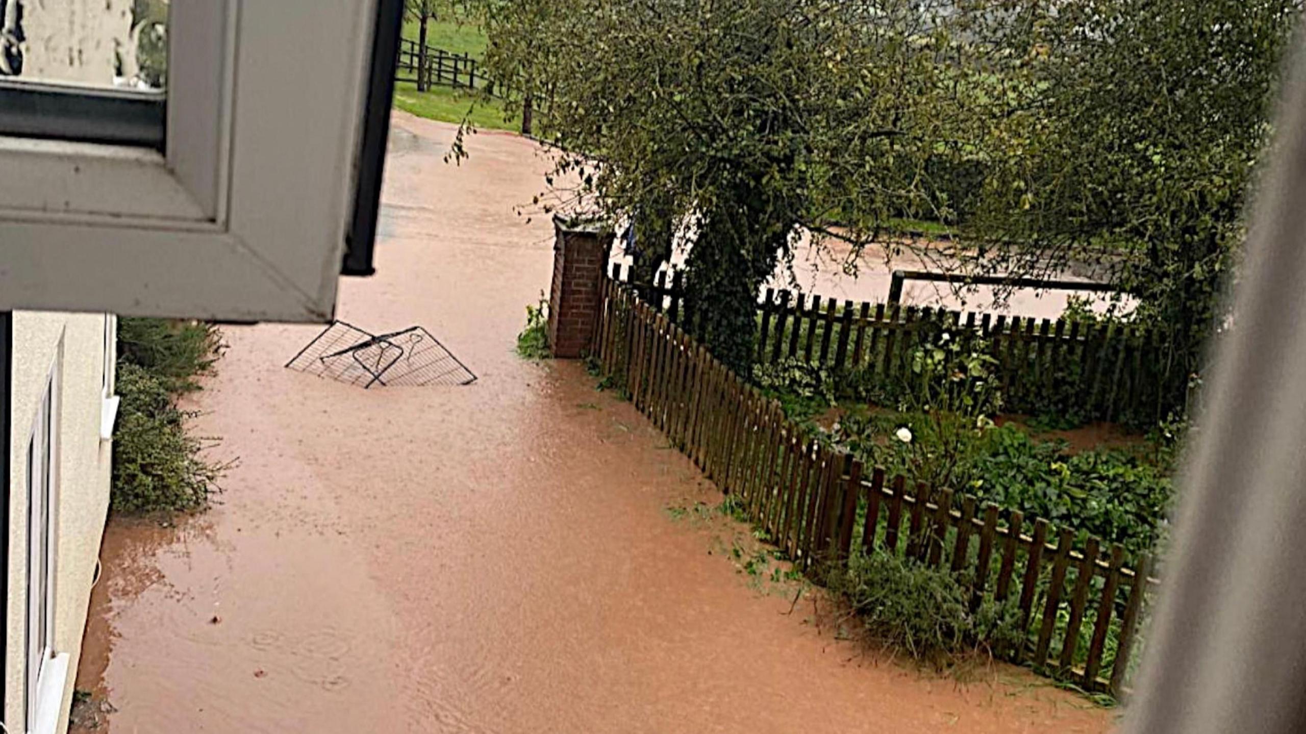 The scene looking out from a bedroom window towards the front of a house where the driveway and the road are covered in brown flood water during Storm Babet in October 2023.