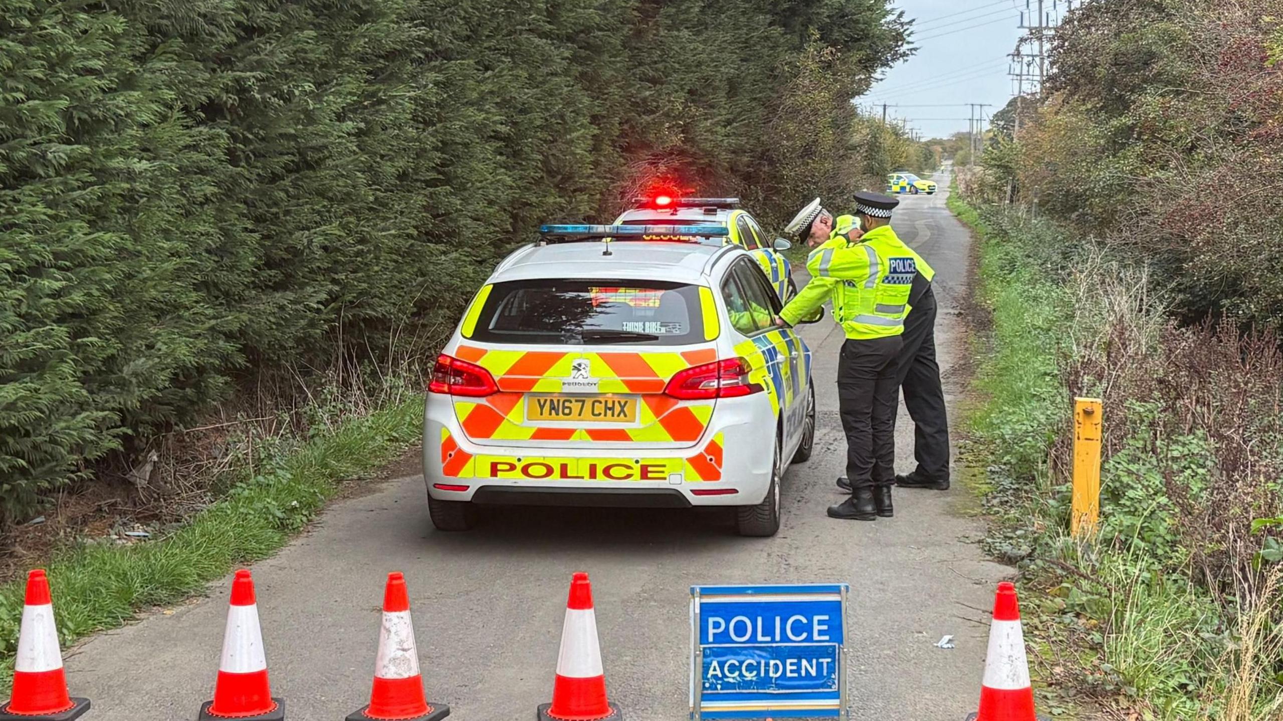 Three police officers wearing yellow hi-vis jackets stand beside two police cars in a narrow country lane, with tall green hedges to either side. In front of the car, the road is blocked by a row of red and white cones and a blue sign with text reading: "Police, accident". Another police car can be seen in the distance.