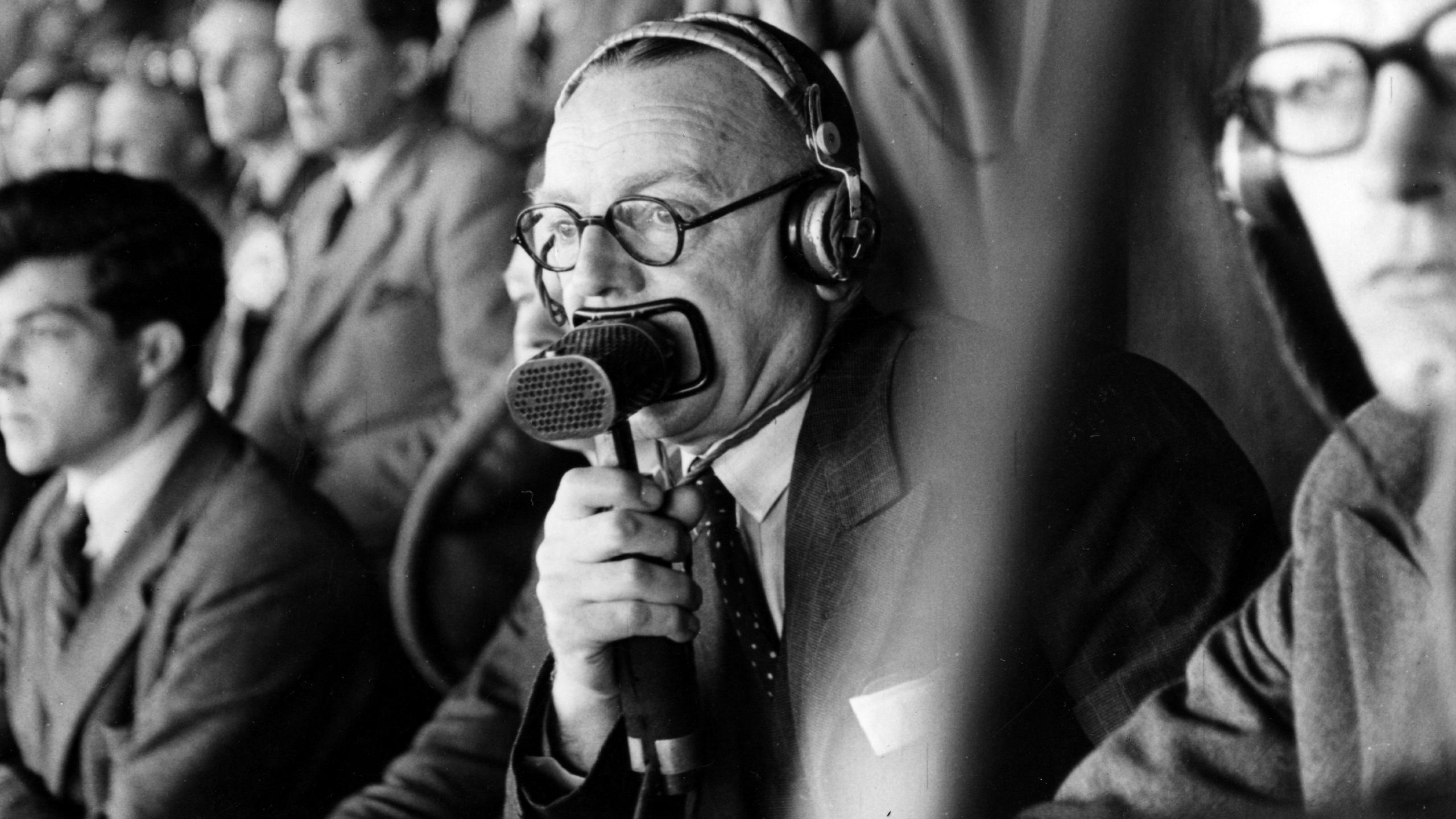 A black-and-white mid-shot of a balding man in round glasses, wearing headphones and with a lip microphone pressed closely against his mouth. He is holding the microphone in his right hand and looking down on events out of frame to the bottom-left. Other people are sitting closely all around him in a crowd, on raked rows of seating going up behind him.