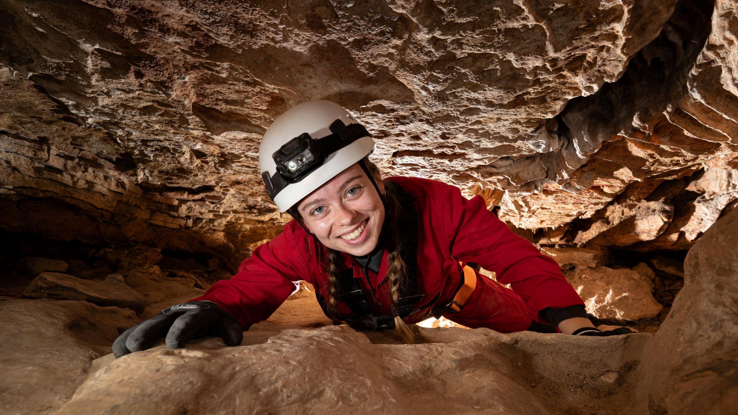 A young woman wearing a white safety helmet, red overalls and black gloves smiles at the camera as she climbs through a narrow passage in the caves at Wookey Hole in Somerset