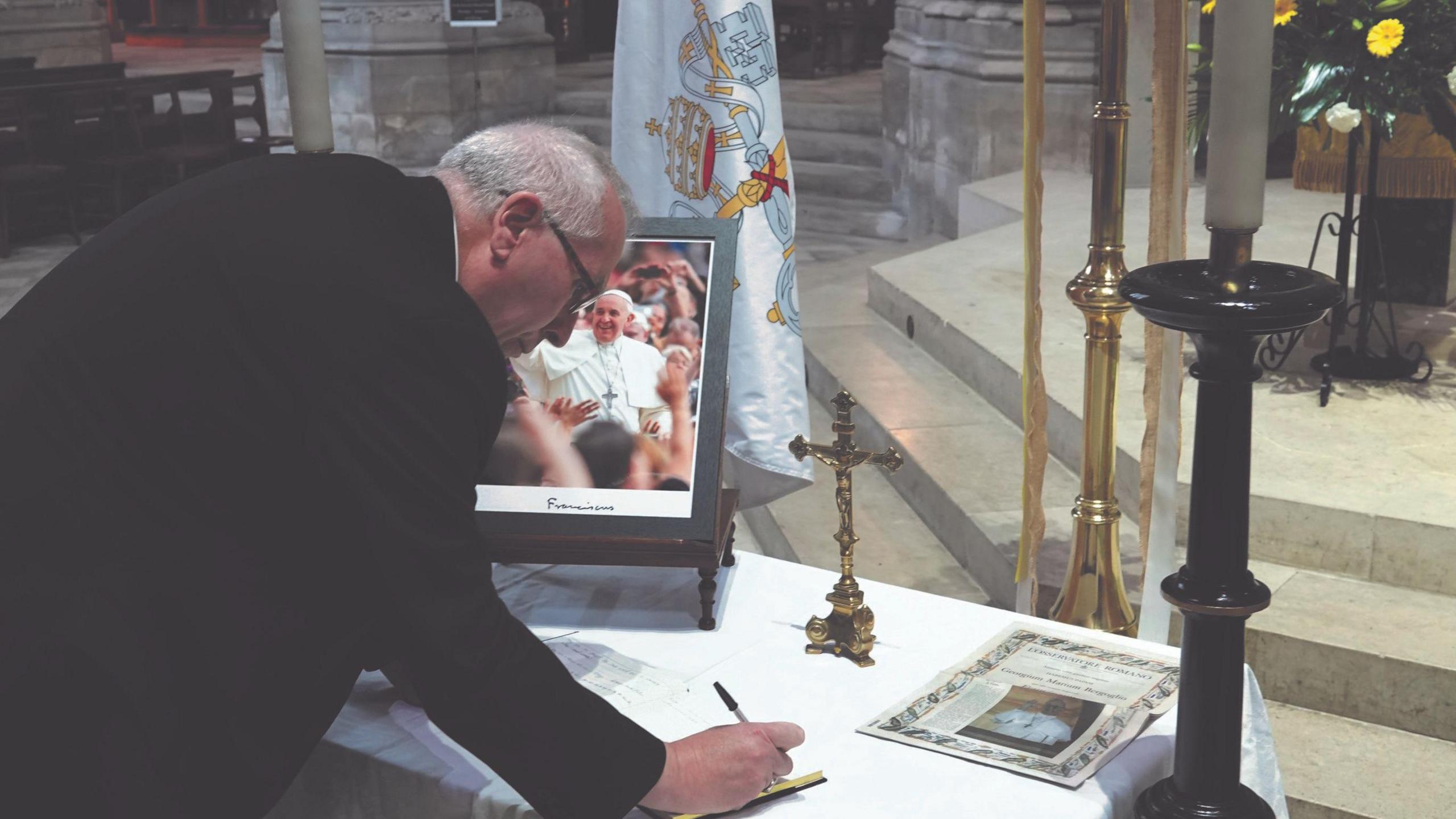 Catholic Bishop of East Anglia, Peter Collins is seen signing a book. He is wearing all black and is signing a book on a table with a white tablet cloth and a framed picture of Pope Francis.