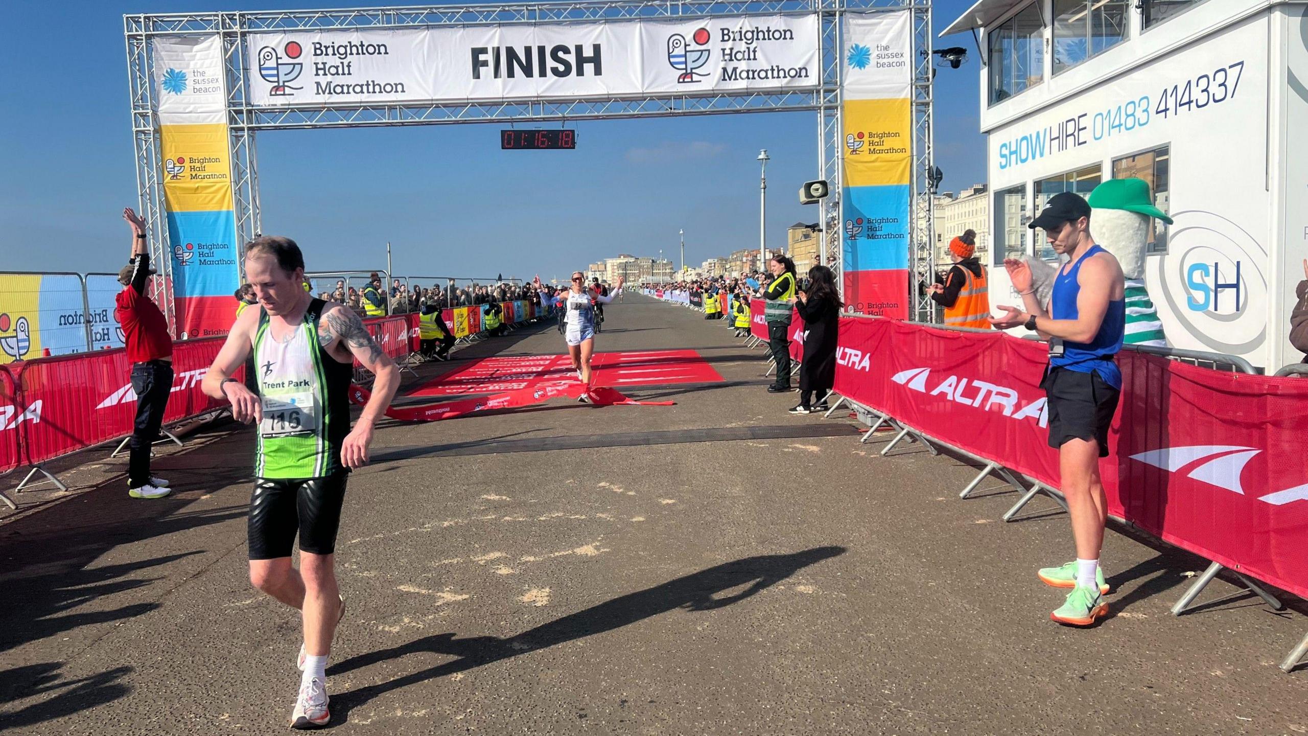 Two runners crossing the finishing line in the 2025 Brighton Half-Marathon, as another runner applauds them.