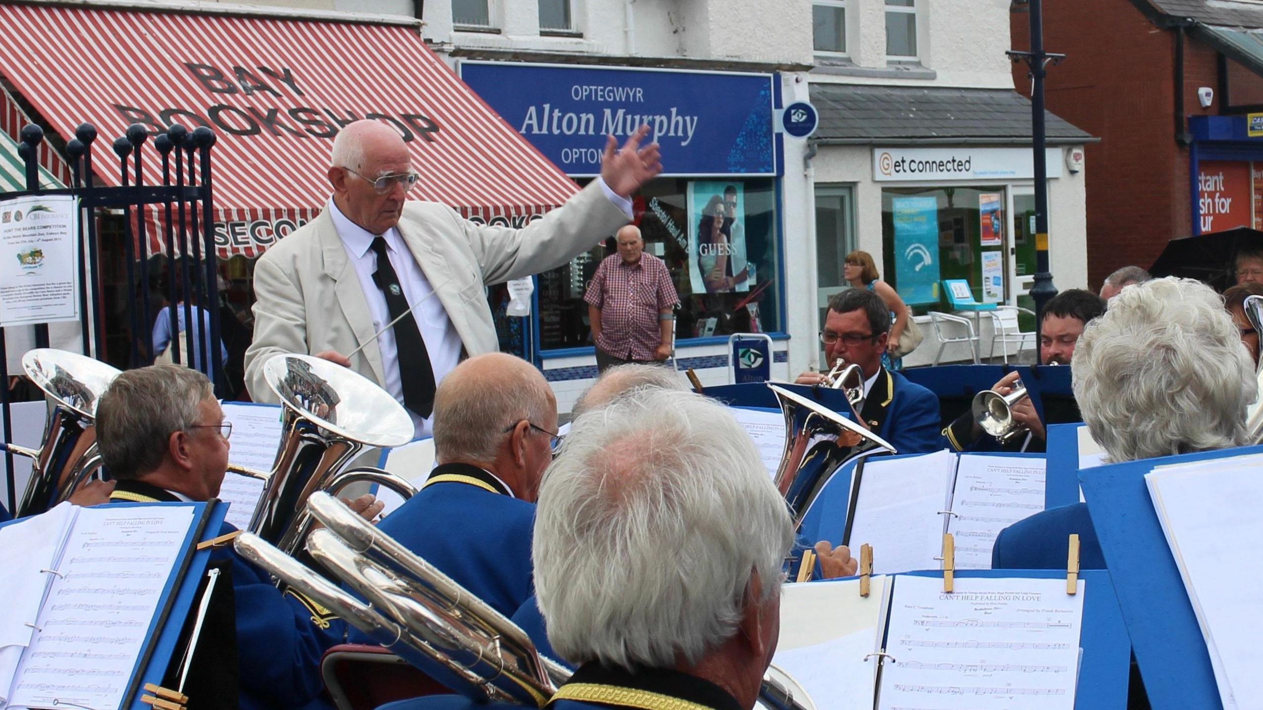 An older man wearing a pale jacket and tie conducts a brass band. Shops are in the background.