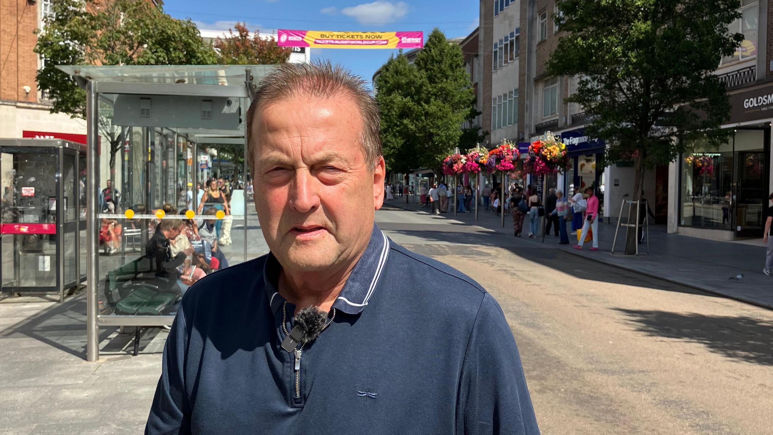 A middle aged man in a blue polo shirt stands in a busy shopping street in the centre of Exeter.