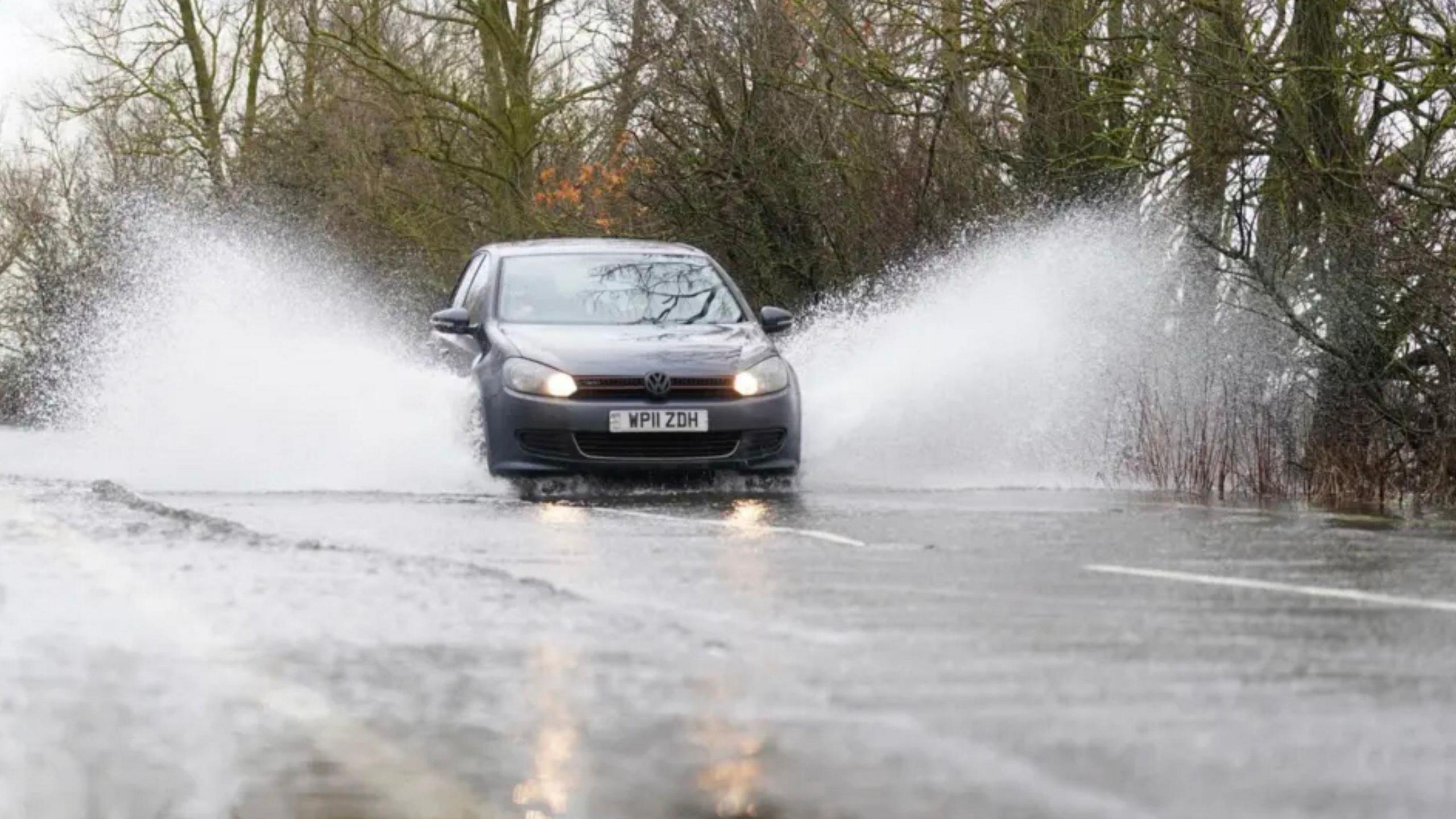 A dark grey sedan car with full beams on sprays standing water either side of it as it goes down a wet, tree-lined road.