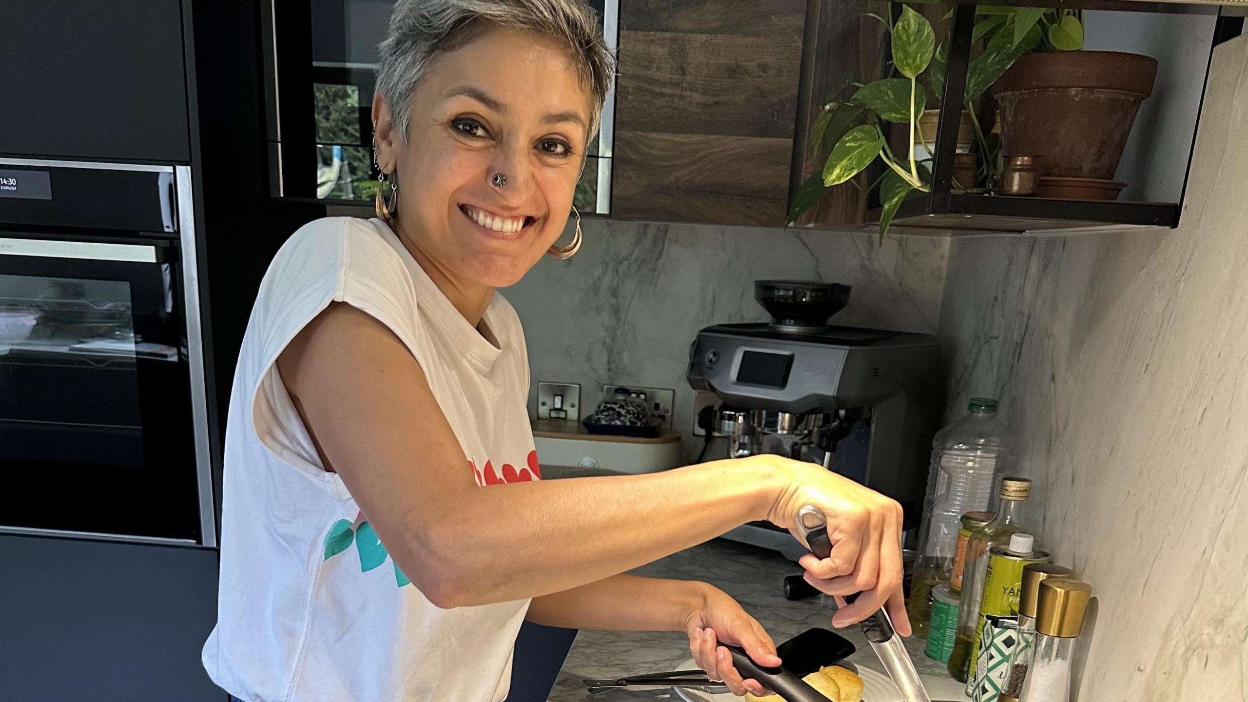 A woman standing in a kitchen holding the handle of a frying pan in one hand and a spatula in the other. She is smiling at the camera and wearing a white t-shirt with green and red hearts on it. In the background there is a coffee machine, a salt a and pepper grinder, some oil, and a plant in a pot on a shelf