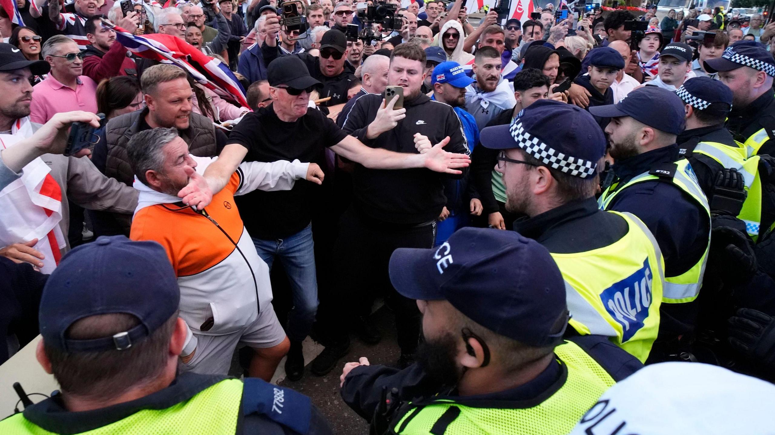 A line of police officers on the right of the picture are faced with a group of protesters, several of who are carrying flags and signs. One man stands holding his phone towards the confrontation